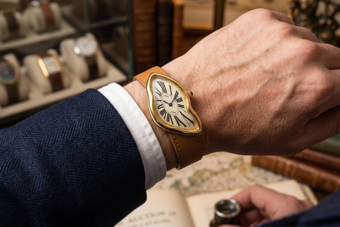 Mavi tüvit ceketli bir bilekte, çarpık ve erimiş görünümlü altın kasalı ikonik Cartier Crash saati; arka planda Sotheby's müzayede evini anımsatan bulanık antika kitaplar ve detaylı bir kataloğun bulunduğu bir masada, eşsiz tasarımının rekor kıran değerini vurgularcasına sergileniyor.