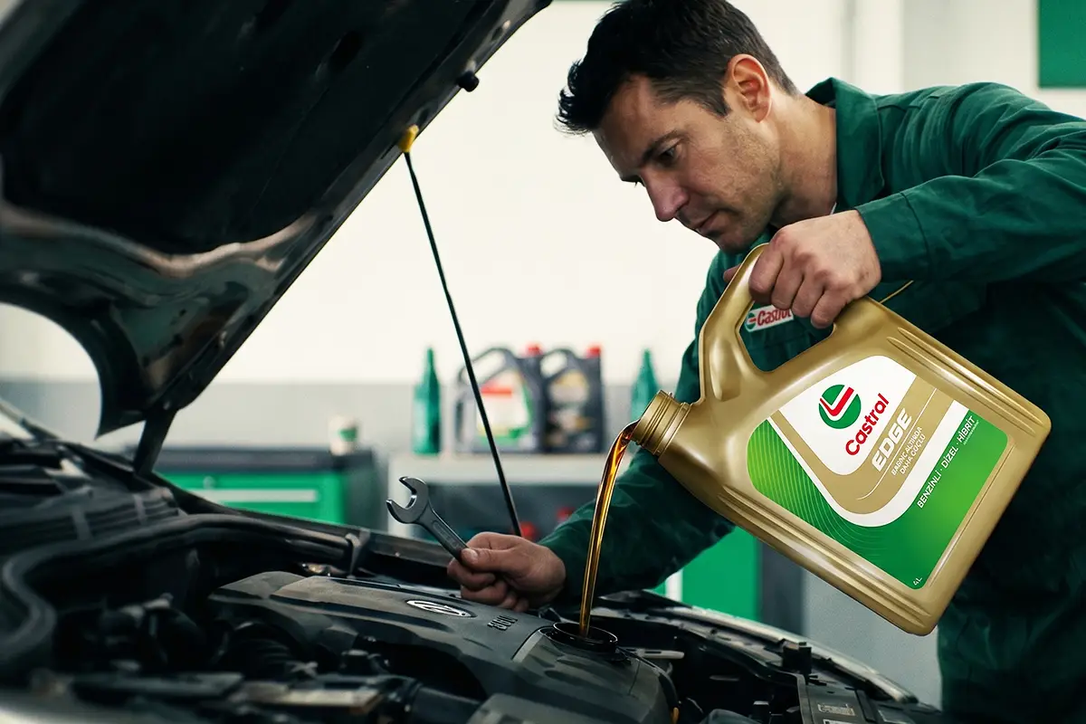 Yeşil üniformalı bir teknisyen, açık kaputlu bir aracın motoruna, üzerinde Castrol EDGE yazan altın renkli bidondan dikkatlice motor yağı eklerken, sol elinde bir anahtar tutarak titiz bir motor bakımı işlemi yapıyor.