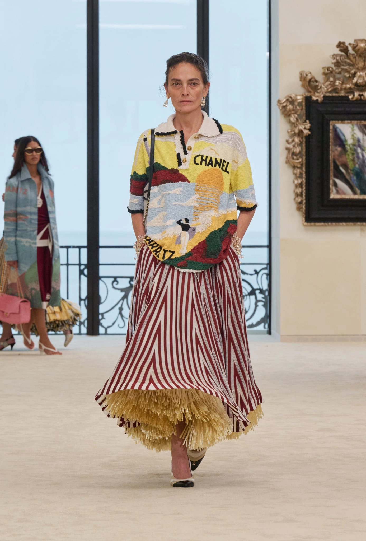 Matthieu Blazy'nin Chanel için Biarritz'de tanıttığı ilk Cruise koleksiyonundan bir model, üzerinde Biarritz yazısı ve sörf yapan insan figürü bulunan sarı, kırmızı, mavi ve yeşil desenli polo yaka triko ile kırmızı beyaz çizgili bir etek giyerek podyumda yürüyor, eteğin altından püsküllü altın rengi detaylar beliriyor.