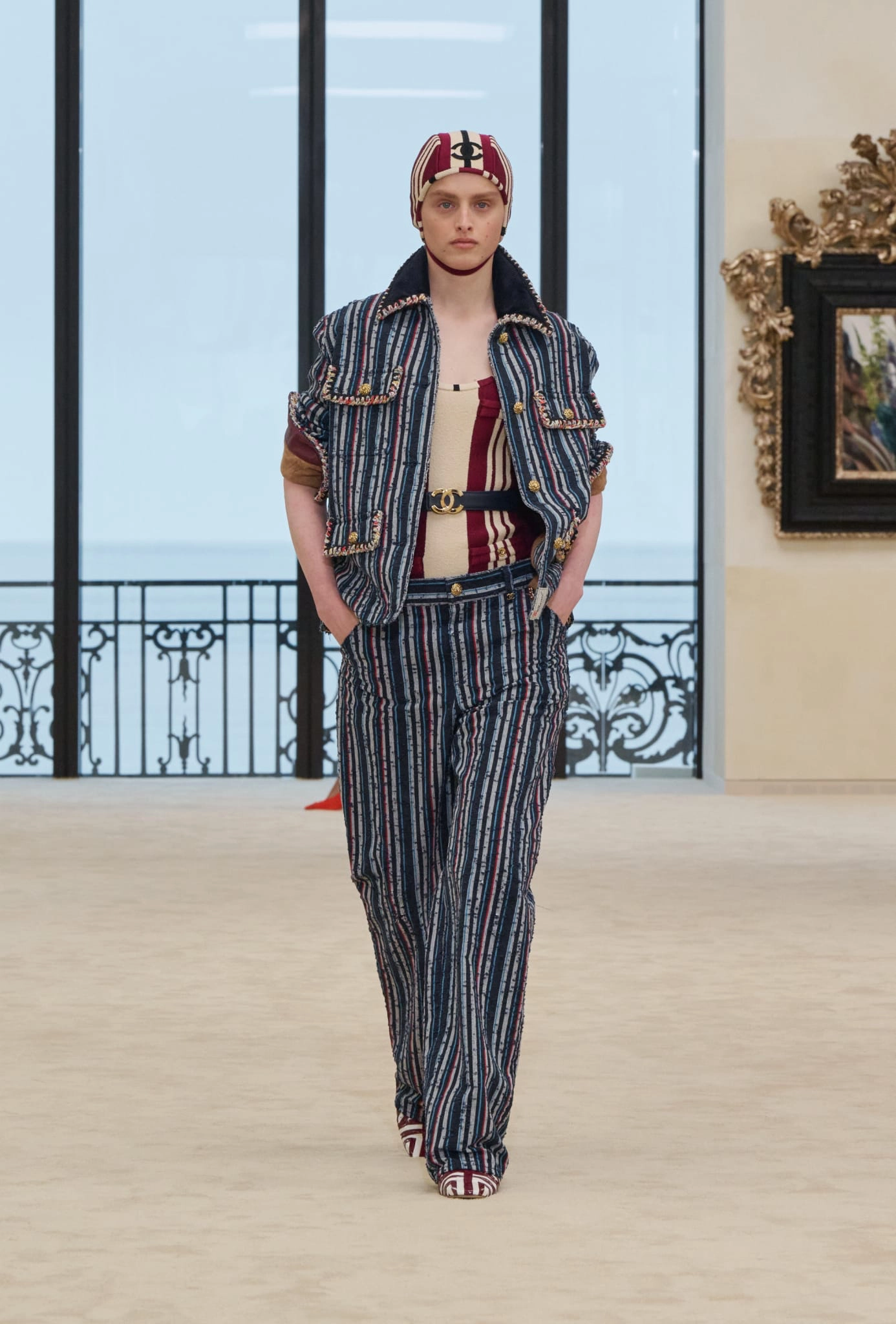 Model, Chanel'in Biarritz'deki ilk Cruise koleksiyonundan, çizgili denizci şapkası ve uyumlu ceket-pantolon takımıyla rahat ve sportif bir zarafeti sergilerken, arka planda okyanus manzarasına açılan pencerelerle koleksiyonun ilham kaynağına gönderme yapıyor.