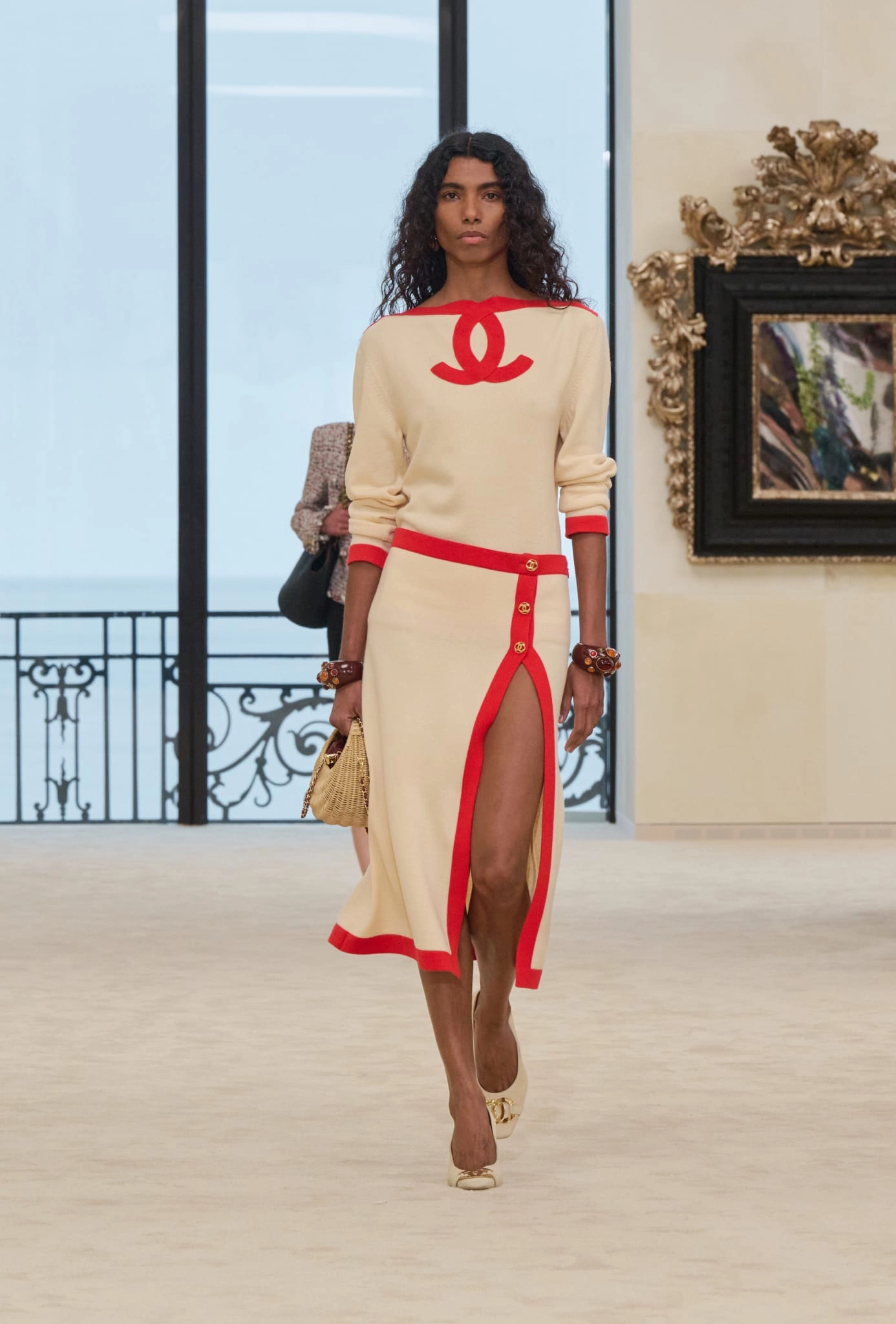 Krem rengi, kırmızı şeritli ve göğsünde büyük kırmızı Chanel logosu bulunan elbisesiyle uzun kıvırcık saçlı bir model, Biarritz'deki ilk Chanel Cruise koleksiyonu defilesinde podyumda yürüyor, arkada okyanus manzaralı pencere ve süslü bir tablo var.