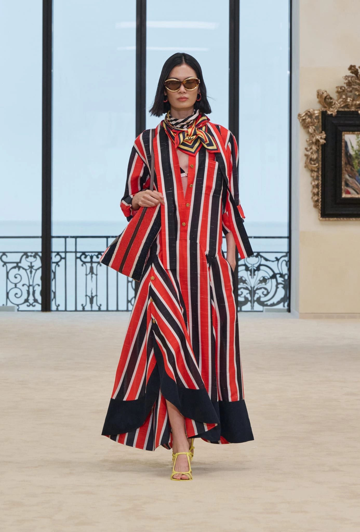 Matthieu Blazy'nin Biarritz'de tanıtılan ilk Chanel Cruise koleksiyonundan, Gabrielle Chanel'in hareket özgürlüğünü ve rahat kesimi yansıtan, dikey kırmızı, siyah ve beyaz çizgili uzun bir elbiseyi, uyumlu ipek fular, çanta ve sarı topuklu ayakkabılarıyla okyanus manzaralı bir podyumda sergileyen bir model.