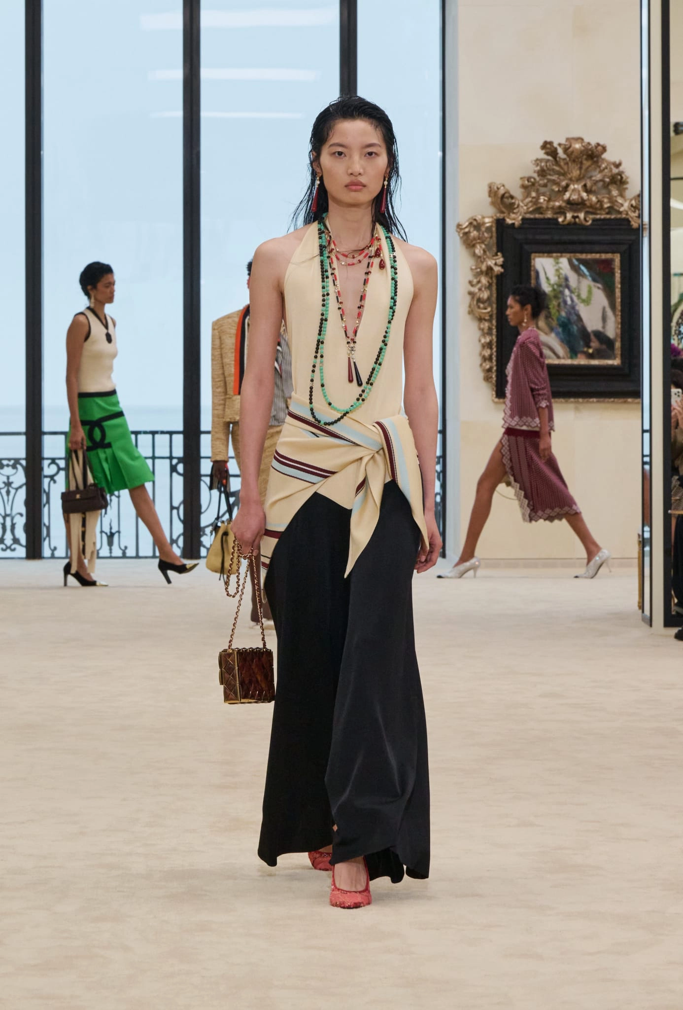 Biarritz'deki ilk Chanel Cruise koleksiyonu defilesinde, krem rengi halter yaka üst, siyah bol paça pantolon ve beline bağladığı desenli kumaşla podyumda yürüyen model, okyanustan ilham alan rahat kesimleri ve özgür ruhu yansıtan çok katmanlı boncuklu kolyeleriyle dikkat çekiyor; arka planda deniz manzarası ve diğer modeller görülüyor.