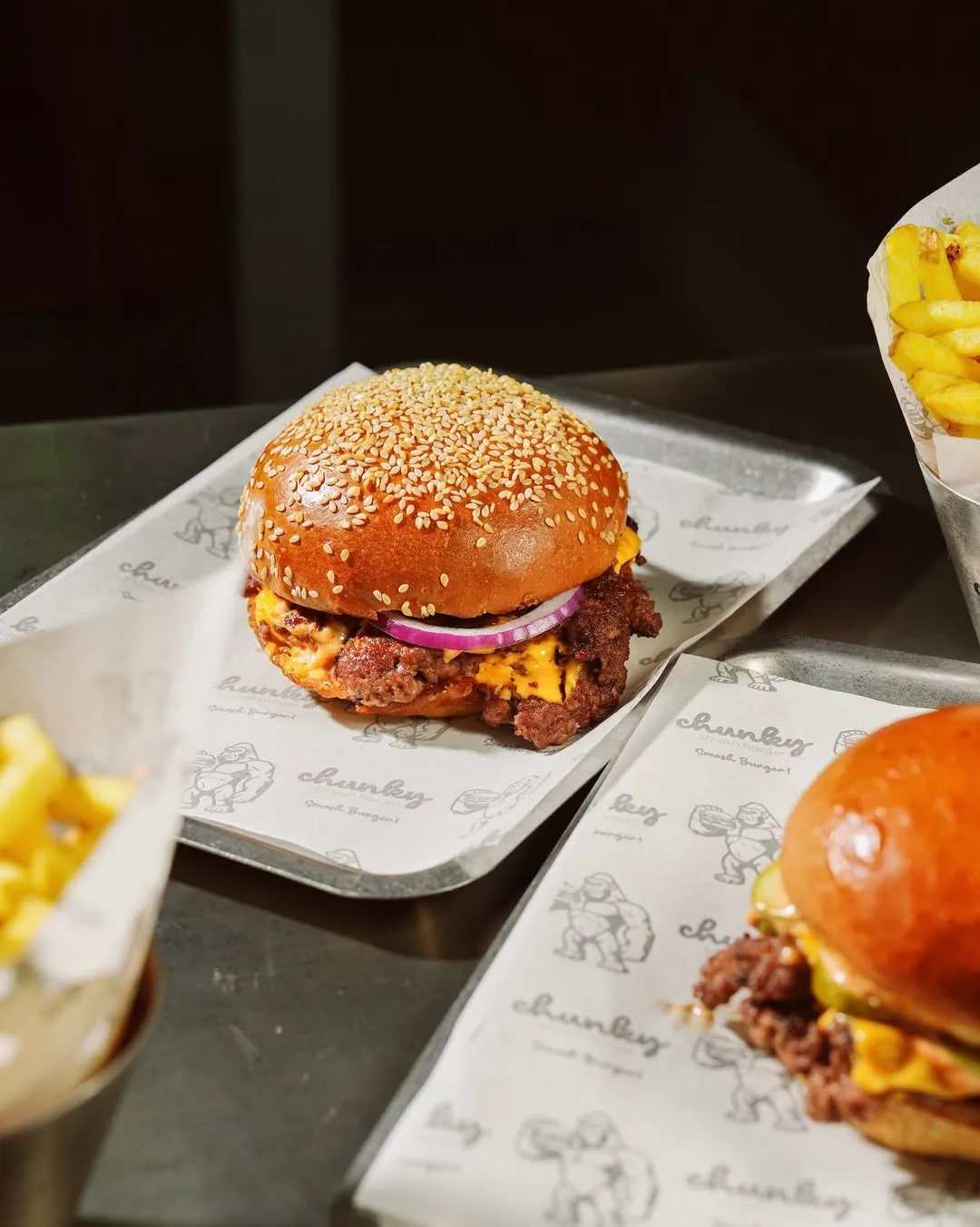 Bir metal tepside, üzerinde susam taneleri olan parlak ekmeği, kalın köftesi, erimiş çedar peyniri ve taze kırmızı soğan dilimleriyle iştah açıcı görünen bir chunky smash burger dikkat çekiyor; arka planda bulanık duran patates kızartmaları ve kısmen görünen başka bir burger ile birlikte, hepsi Chunky Smash Burger logolu özel ambalaj kağıtlarına sarılı.