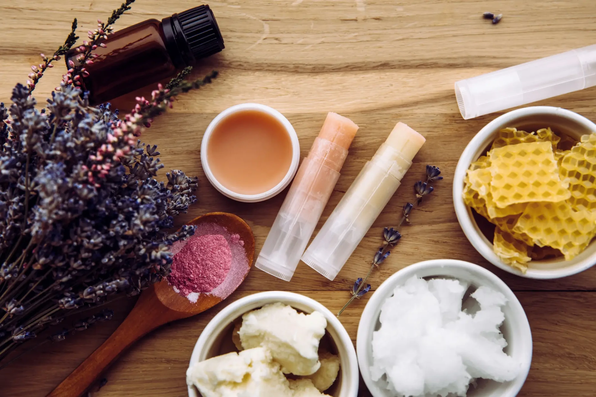 Ahşap bir yüzey üzerinde, doğal içerikli lip balm yapımında kullanılan lavanta demeti, bal mumu parçaları, shea yağı ve Hindistan cevizi yağı gibi malzemelerin yanı sıra şeffaf ambalajlarda ve küçük bir kapta hazırlanmış farklı tonlarda lip balmlar düzenli bir şekilde sergileniyor.