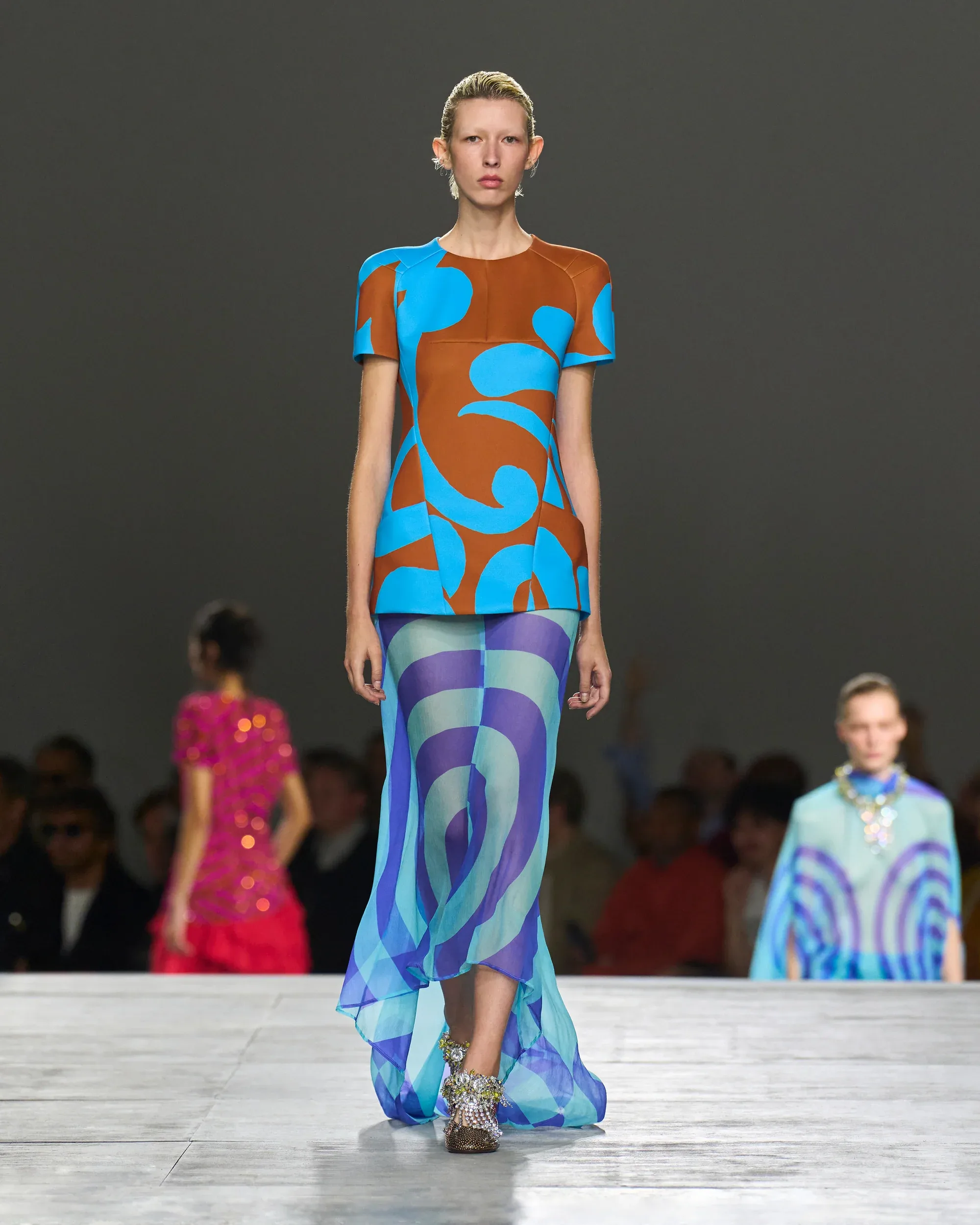 Dries Van Noten'in 2026 ilkbahar-yaz moda trendlerini sergilediği podyumda, kahverengi ve turkuazın güçlü desenlerini taşıyan kısa kollu bluzu ile mavi-mor tonlarında dalgalı, şeffaf, hareketli bir etek giyen model, sezonun canlı renk ve dokusal vurgusunu gözler önüne seriyor.