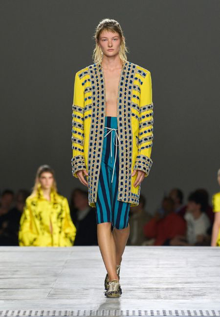 Dries Van Noten'in 2026 İlkbahar-Yaz koleksiyonundan canlı sarı, lacivert ve bej işlemeli geometrik desenlerle süslenmiş, dikkat çekici güçlü bir silüete sahip açık ceketi ve turkuaz-lacivert dikey çizgili şortlarıyla podyumda yürüyen bir model, sezonun öne çıkan dokulu yüzey ve canlı renk trendlerini yansıtıyor.