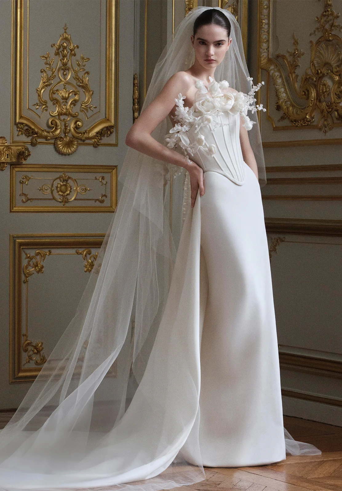 Elie Saab 2027 İlkbahar gelinlik koleksiyonundan, straplez, korse detaylı ve göğüs ile omuzlarında üç boyutlu fildişi çiçek işlemeleriyle zenginleştirilmiş modern kesim bir gelinlik giyen model, arkasındaki gösterişli duvar ve uzun duvağıyla zarif bir duruş sergiliyor.