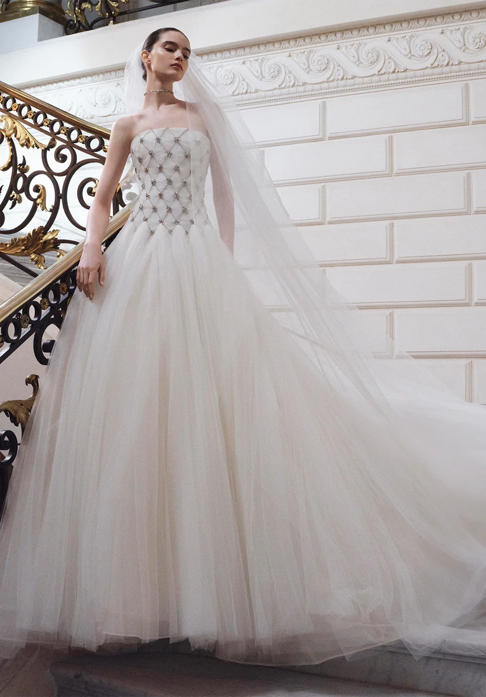 Elie Saab 2027 gelinlik koleksiyonundan, bedeni pırlanta desenli işlemelerle süslü straplez korseli ve kabarık tül etekli, uzun duvağı yere serilmiş zarif bir gelin, oymalı altın varaklı merdivenli şık bir salonda düşünceli bir ifadeyle duruyor.