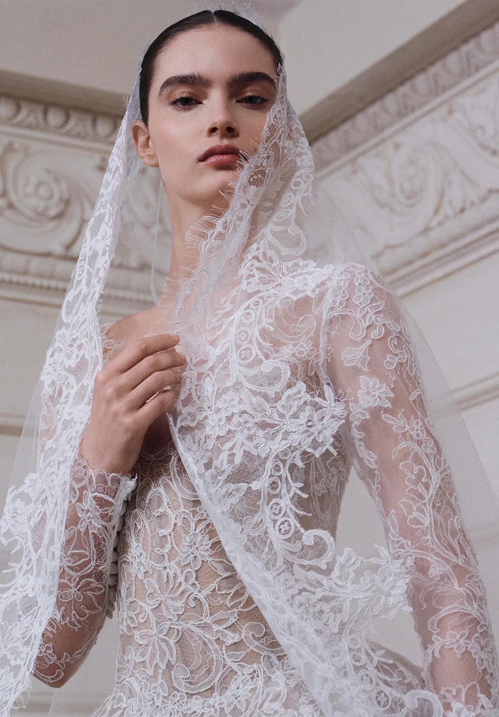 Elie Saab 2027 İlkbahar gelinlik koleksiyonundan, zarif çiçek desenli şeffaf dantel bir elbise ve narin işlemeli duvakla süslenmiş model, Paris saraylarının ihtişamını anımsatan duvar detayları önünde doğrudan izleyiciye bakarak romantik ve zamansız bir gelinlik vizyonunu sergiliyor.
