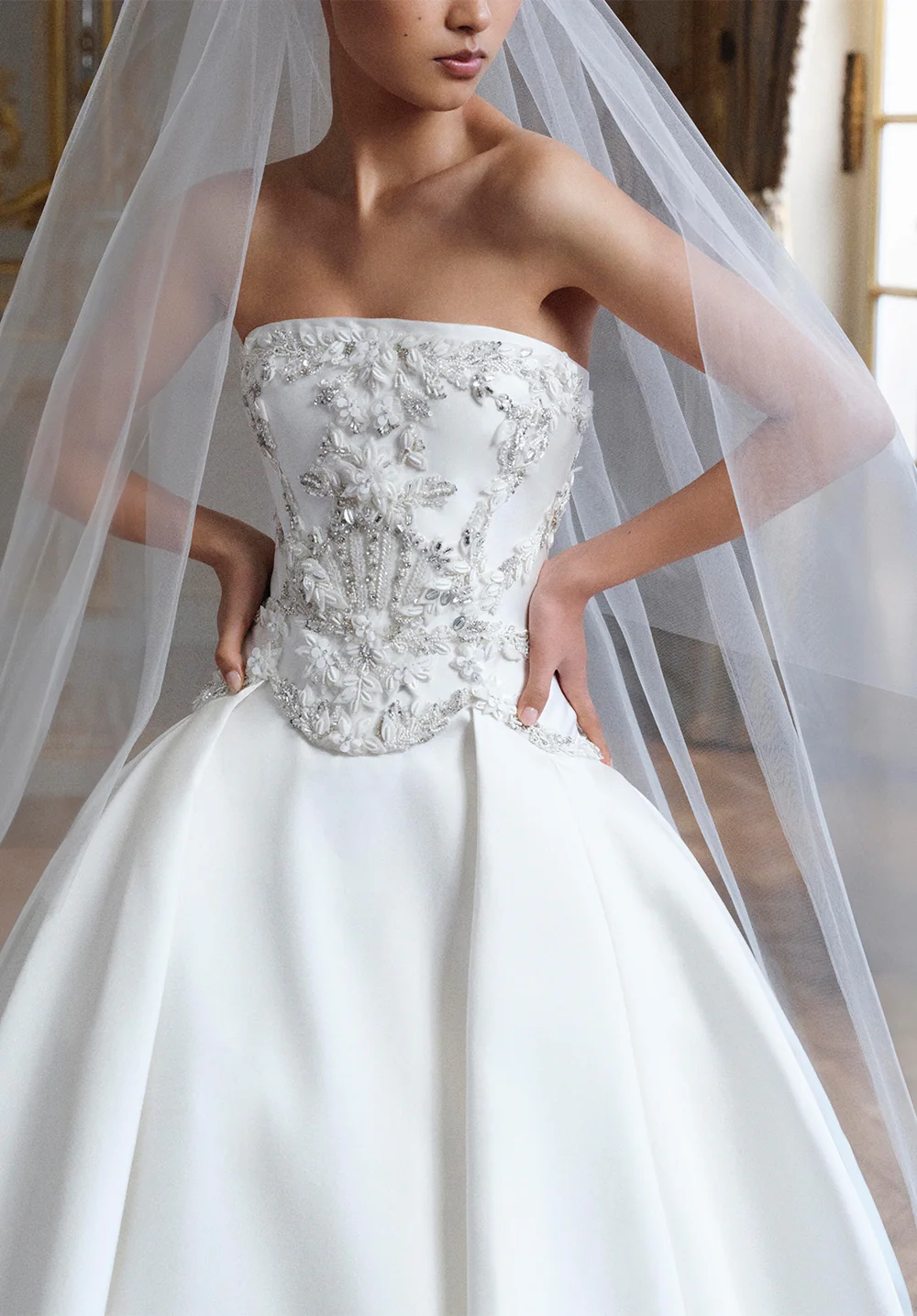 Elie Saab 2027 İlkbahar gelinlik koleksiyonundan, fildişi çiçek nakışları, kristaller ve metalik örgü dokularla detaylandırılmış, basque bel kesimli straplez bir korsaj ve kabarık Mikado etekle tamamlanan, uzun tül duvaklı beyaz bir gelinlik içindeki model, Paris sarayı atmosferinde zarafetle poz veriyor.