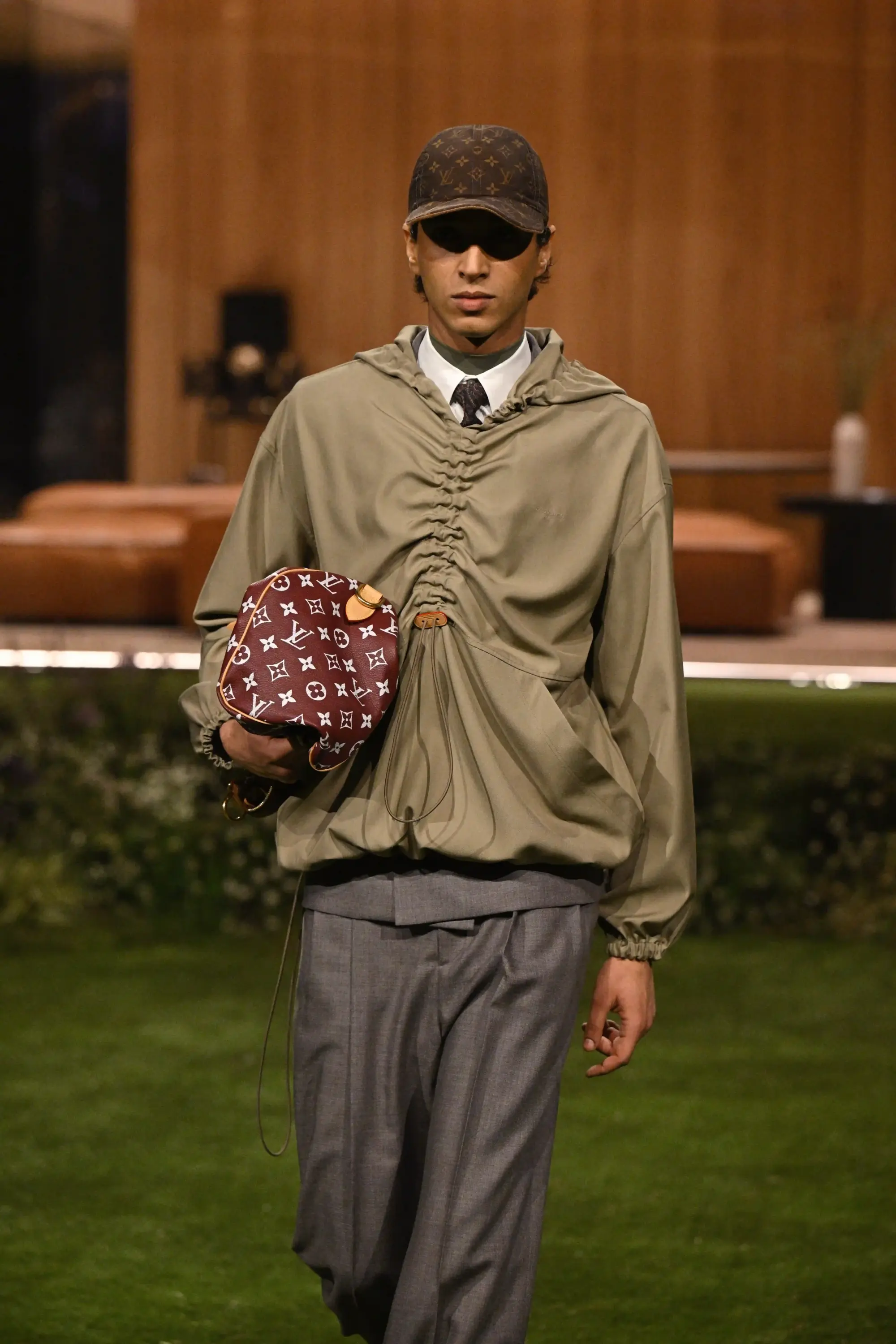 Podyumda yürüyen bir erkek model, haki renkli kapüşonlu ceket, gri pantolon, koyu kahverengi Louis Vuitton monogram desenli şapka ve bordo renkli Louis Vuitton monogram desenli yuvarlak bir çanta taşıyor, bu görünüm erkeklerde renkli aksesuar kullanımının stil üzerindeki etkisini vurguluyor.