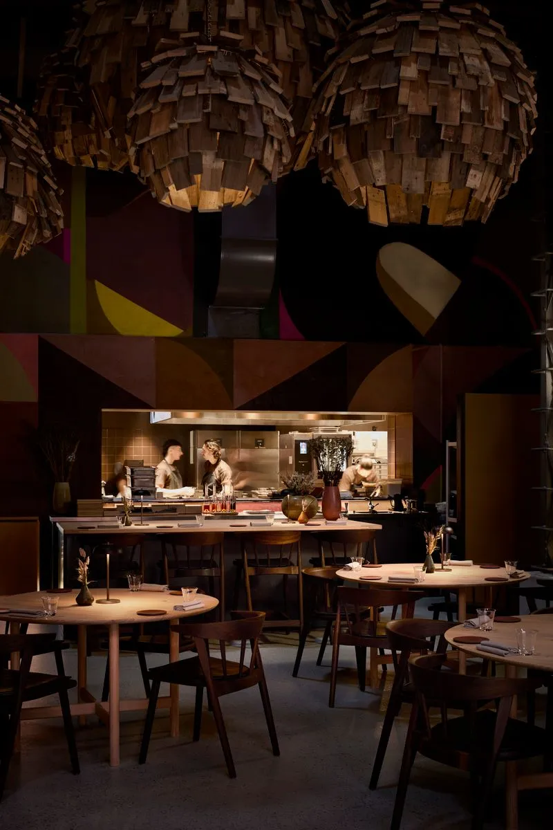 Kopenhag'daki Esse restoranının modern ve samimi iç mekanında, ahşap parçalarla kaplı küresel avizelerin sıcak ışığı altında, hareketli açık mutfakta şefler çalışırken, zarif ahşap masalar ve sandalyeler misafirleri bekliyor.