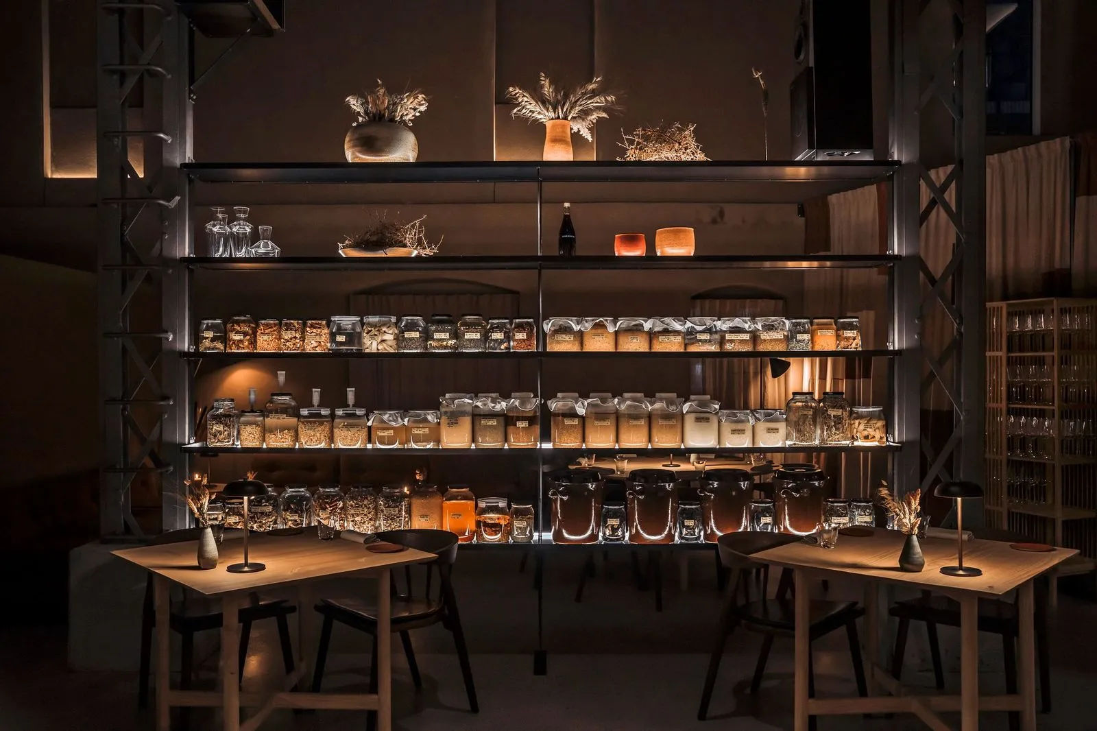 Kopenhag'daki Esse restoranının loş ışıklı iç mekanında, endüstriyel raflarda sıralanmış, içinde çeşitli tahıllar, baharatlar ve sıvıların bulunduğu yüzlerce cam kavanoz dikkat çekiyor; ön planda ise sıcak bir atmosfer sunan iki ahşap masa ve sandalye takımı yer alıyor.