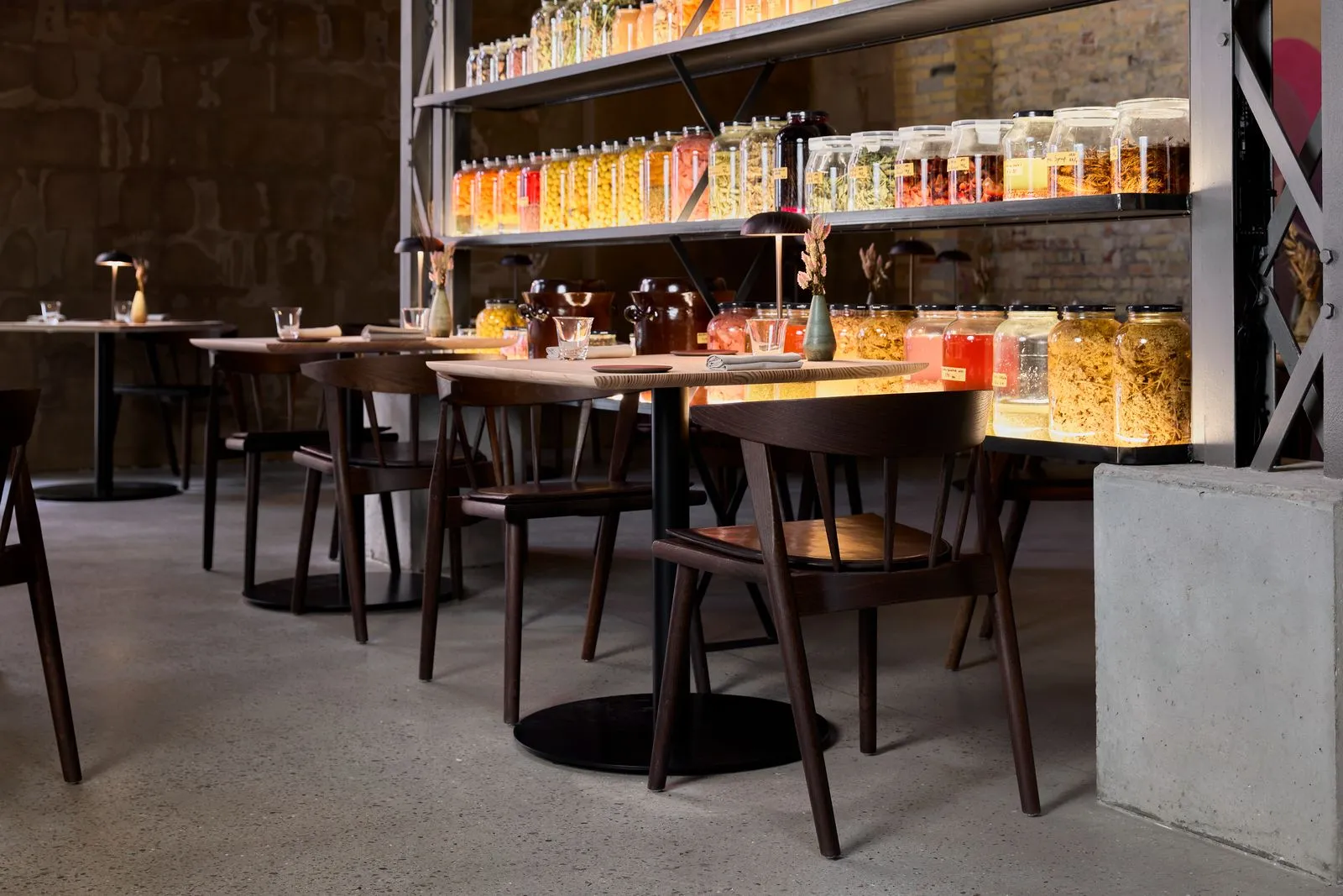 Kopenhag'daki Esse restoranının modern ve sıcak iç mekanında, ahşap masalar ve sandalyeler özenle yerleştirilmiş, arka planda loş ışıklarla aydınlatılmış raflarda dizili rengarenk cam kavanozlar içindeki konserve yiyecekler dikkat çekiyor.