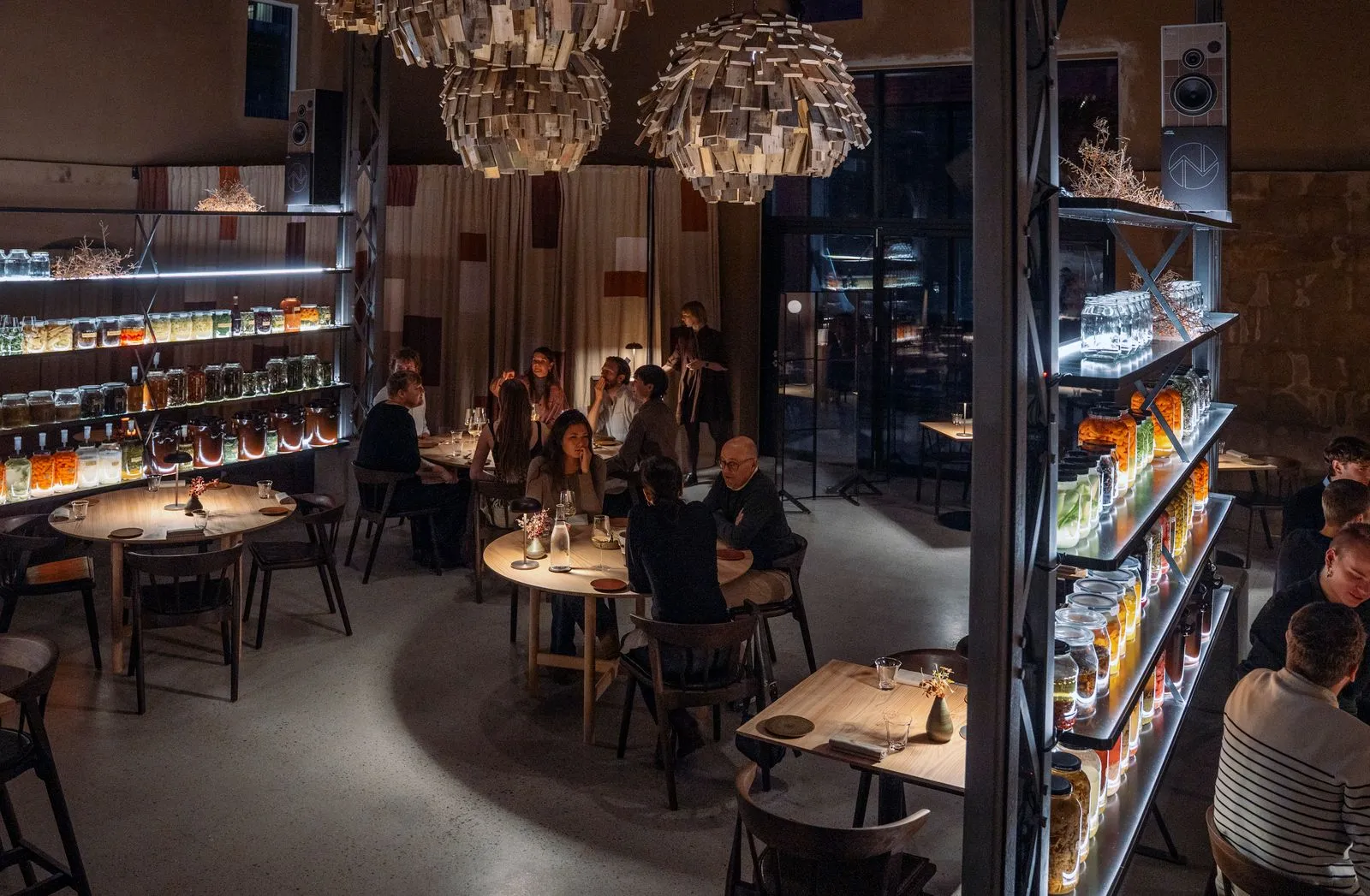 Kopenhag'daki Esse restoranının modern ve loş atmosferinde, ahşap masalarda sohbet edip yemek yiyen misafirlerin arasında, parlak ışıklı raflarda düzenlenmiş renkli cam kavanozlar ve tavandan sarkan özgün tasarımlı küresel lambalar dikkat çekiyor.