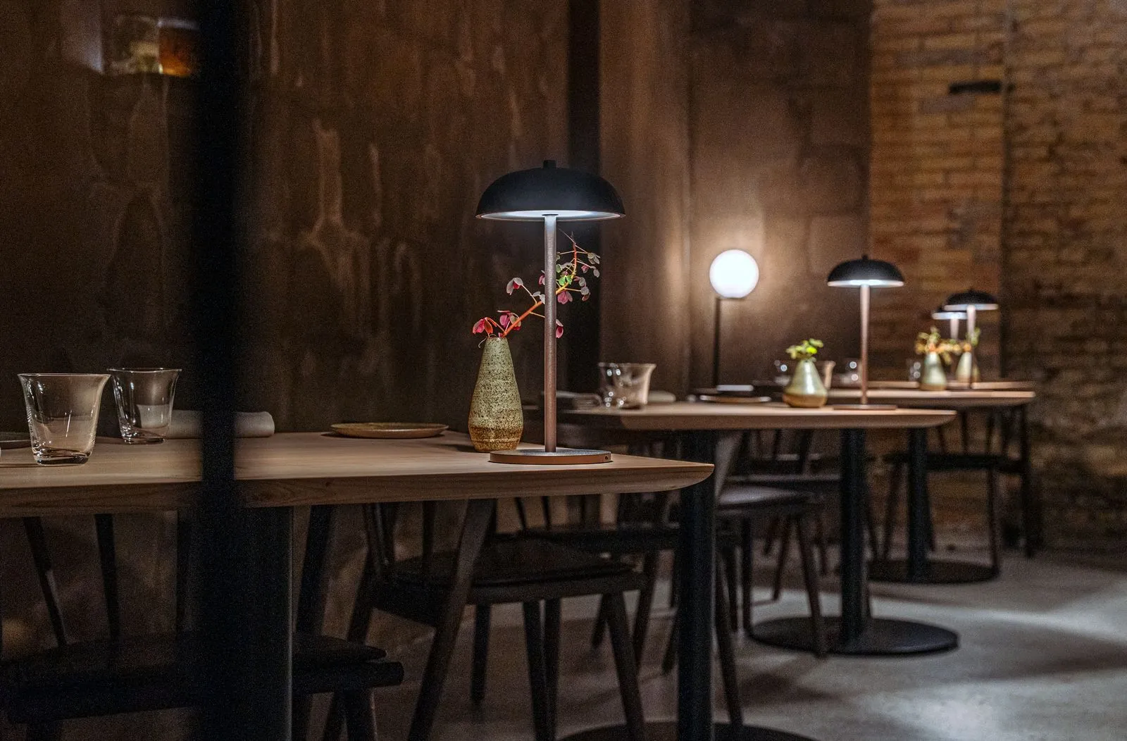 Kopenhag'daki Esse restoranının karakteristik iç mekanında, karanlık duvarlar önünde sıralanmış ahşap masaların üzerinde, loş ışık yayan modern siyah kubbeli masa lambaları ve zarif çiçek vazoları, misafirlere sıcak ve samimi bir yemek deneyimi vadeden sofistike bir atmosfer yaratıyor.
