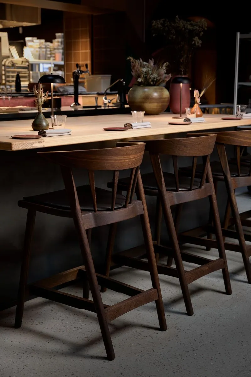 Kopenhag'da bulunan şık bir restoran veya barın iç mekanından, loş ışıklandırmanın hakim olduğu uzun ahşap bar tezgahı ve önüne sıralanmış koyu ahşap bar sandalyelerinin modern ve davetkar bir yemek deneyimi sunduğu görülüyor.