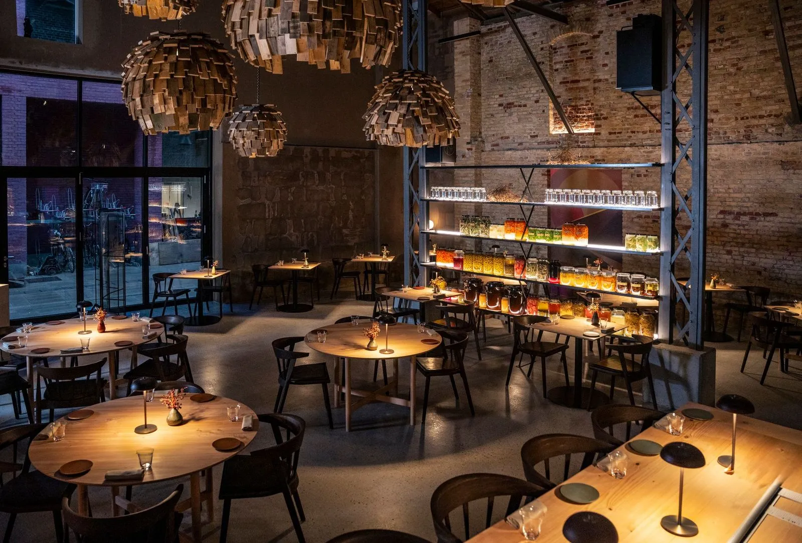 Esse Restaurant'ın geniş iç mekanında, ahşap parçalı şık avizelerin loş ışıklarla aydınlattığı modern ve endüstriyel bir tasarımla döşenmiş, boş ahşap masalar sıralanıyor, duvarlardaki tuğla dokusu ve renkli kavanozların sergilendiği raflar mekana sıcak bir ambiyans ve özel bir yemek deneyimi katıyor.