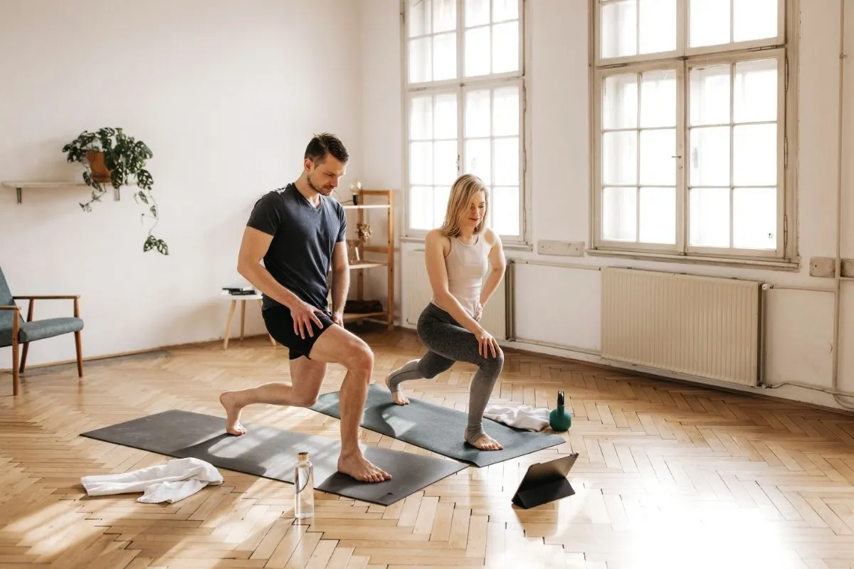 Evde spor yapan bir erkek ve bir kadın, aydınlık bir odada yoga matlarının üzerinde lunge egzersizi gerçekleştirirken, önlerinde su şişesi, havlu, kettlebell ve tablet gibi temel ekipmanlar, etkili bir evde spor programı uygulamak için uygun bir ortam sergiliyor.