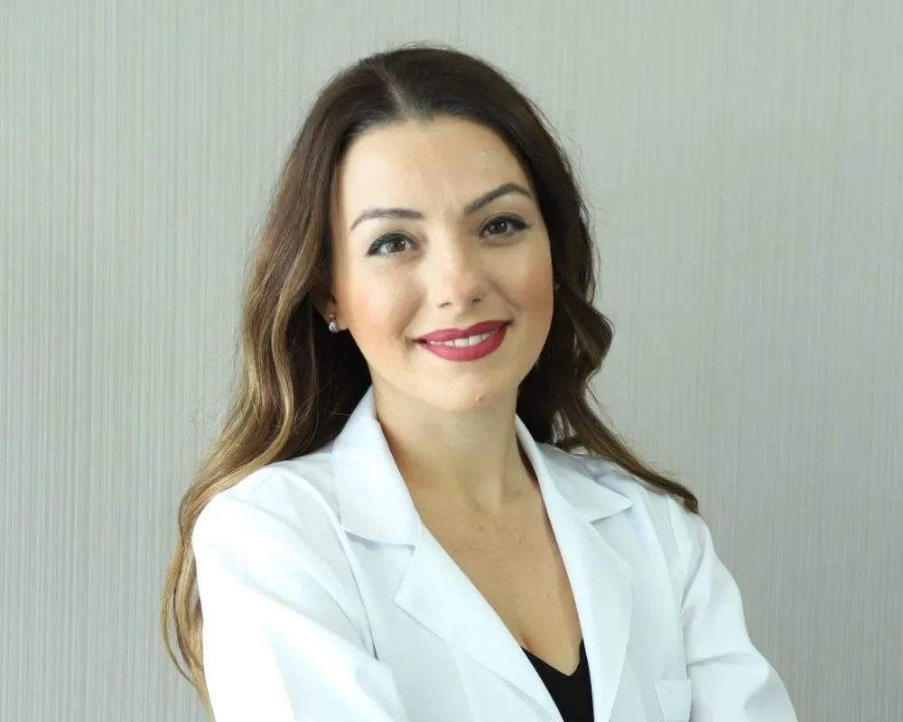 Ezgi Özkur, beyaz önlüğü içinde kameraya zarifçe gülümserken, uzun dalgalı kahverengi saçları, belirgin kırmızı ruju ve parlak küpeleriyle Düğün Öncesi Bakım gibi konulardaki uzmanlığını yansıtan profesyonel bir duruş sergiliyor.