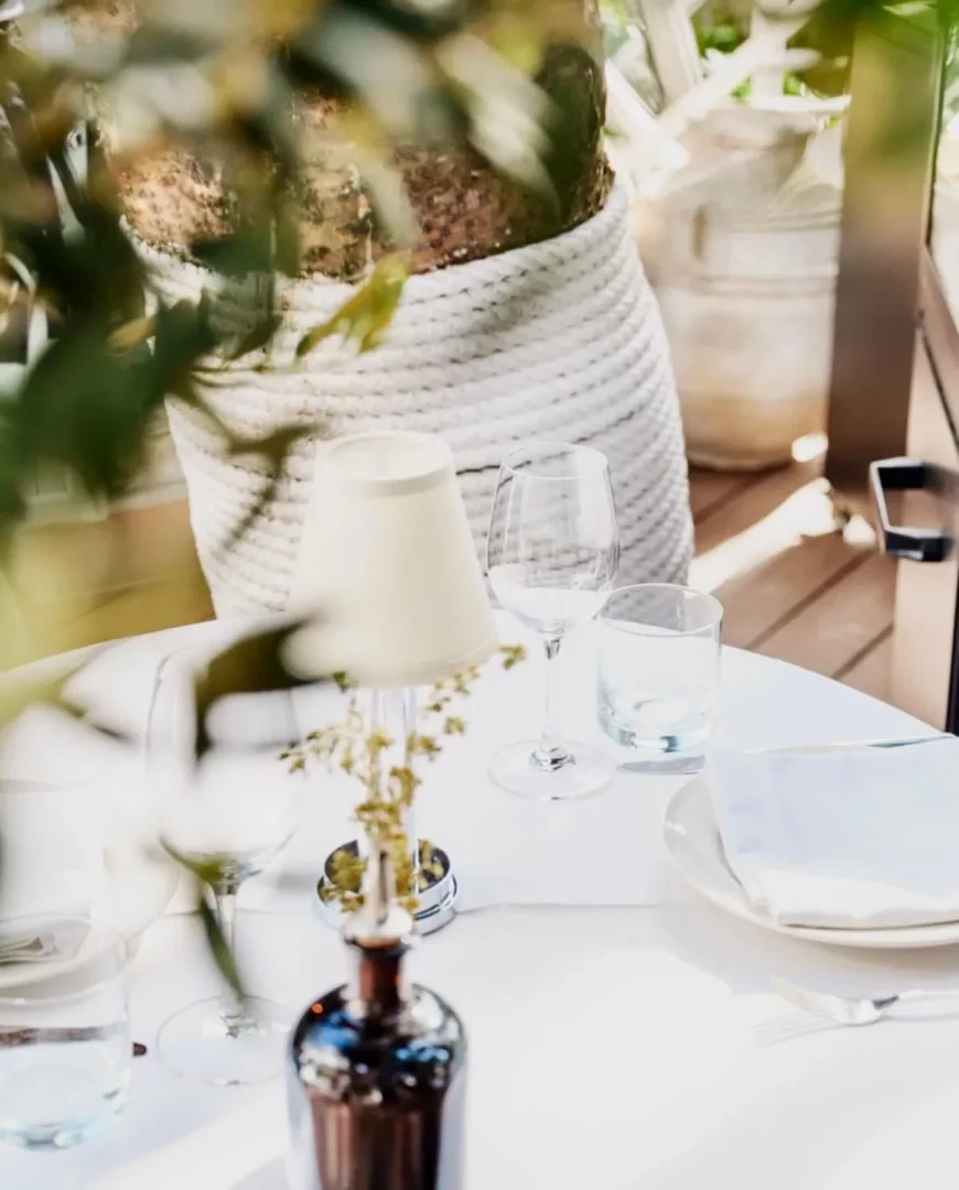 Bodrum'daki bir mekanda, beyaz masa örtülü yuvarlak bir masanın üzerinde şarap ve su kadehleri, tabaklar, peçete ve ortasında şık bir masa lambası ile yeşillik detaylarını gösteren, sıcak bir öğle yemeği veya akşam yemeği atmosferini yansıtan bir görsel.