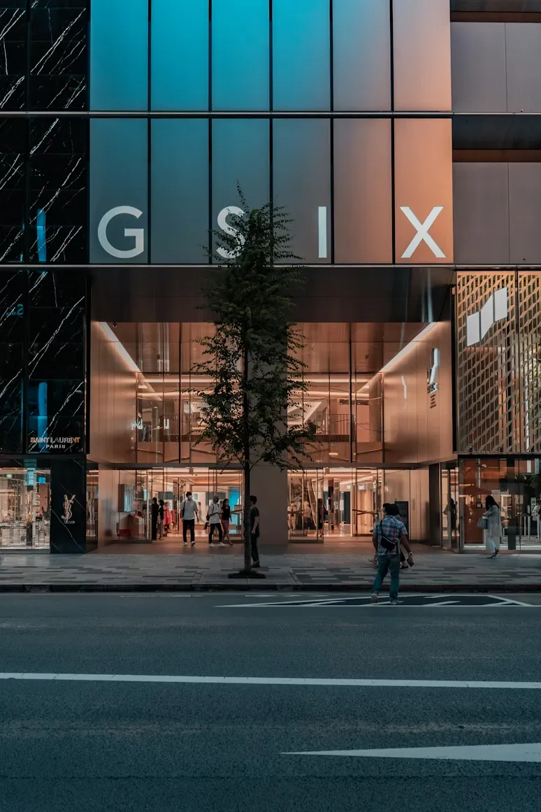Ginza SIX binasının etkileyici modern cam cephesi, üzerinde büyük G SIX yazısı, önünde bir ağaç ve Saint Laurent Paris mağazasının girişiyle birlikte, hareketli Ginza alışveriş bölgesindeki canlı atmosferi yansıtan, hem içeride hem dışarıda insanların olduğu dinamik bir sokak görünümü.