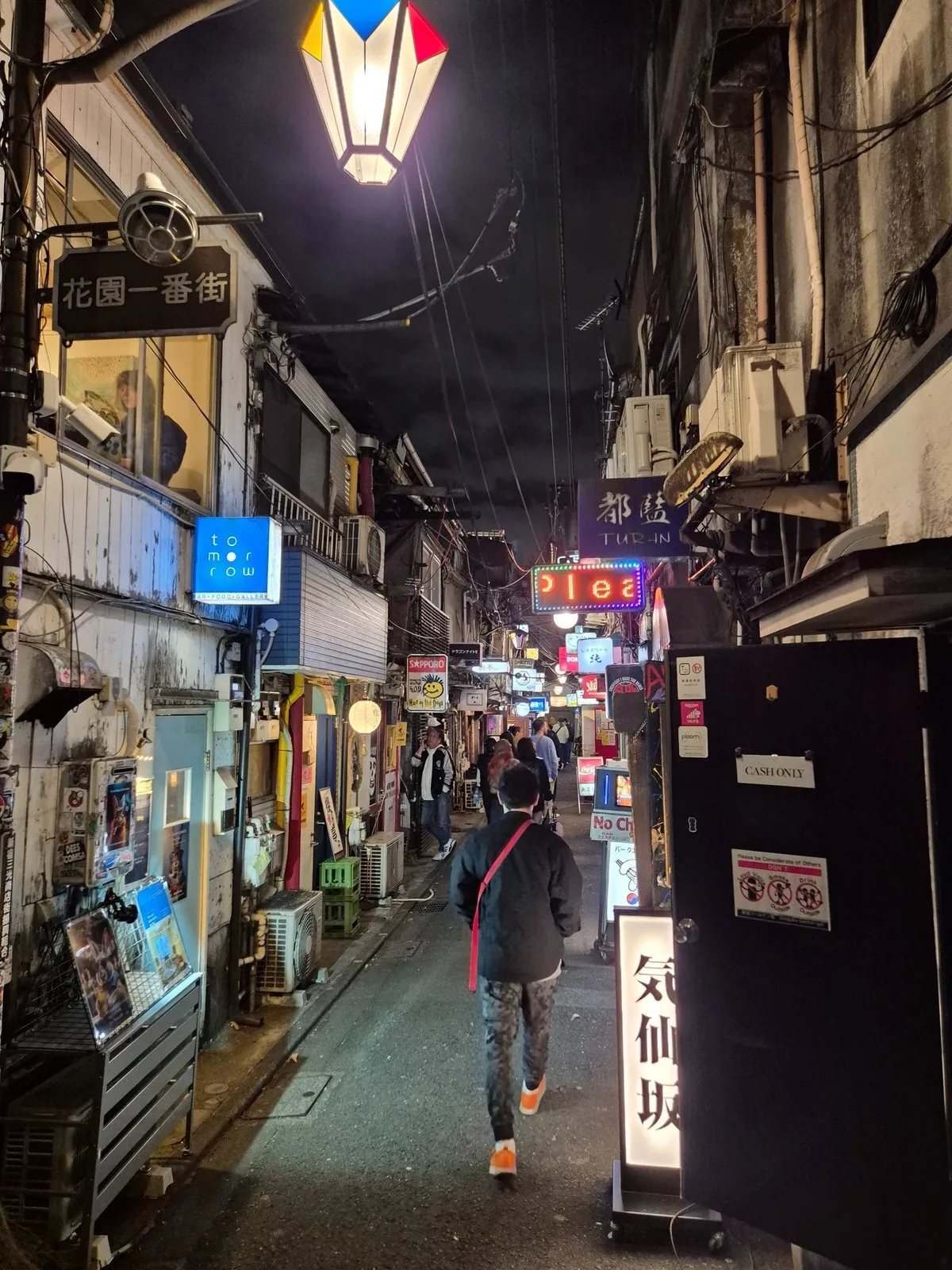 Tokyo'nun ikonik Golden Gai bölgesindeki dar bir sokakta, bir kişinin önünde yürüdüğü, tepeden sarkan rengarenk fenerin ışığı altında yüzlerce küçük barın neon tabelaları ve klimalarla dolu, hareketli ve özgün bir gece manzarası görülüyor.
