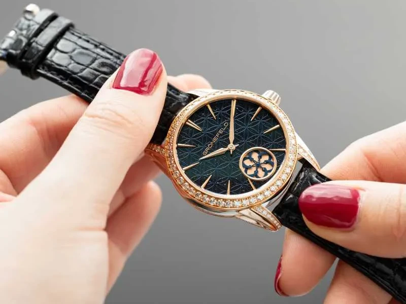 Kırmızı ojeli tırnaklara sahip eller, kadranında zarif geometrik desenler ve alt kısmında şık bir süs motifi bulunan, etrafı parlak elmaslarla bezeli rose gold kasalı Grönefeld Tanfana marka lüks bir kol saatini inceliyor.