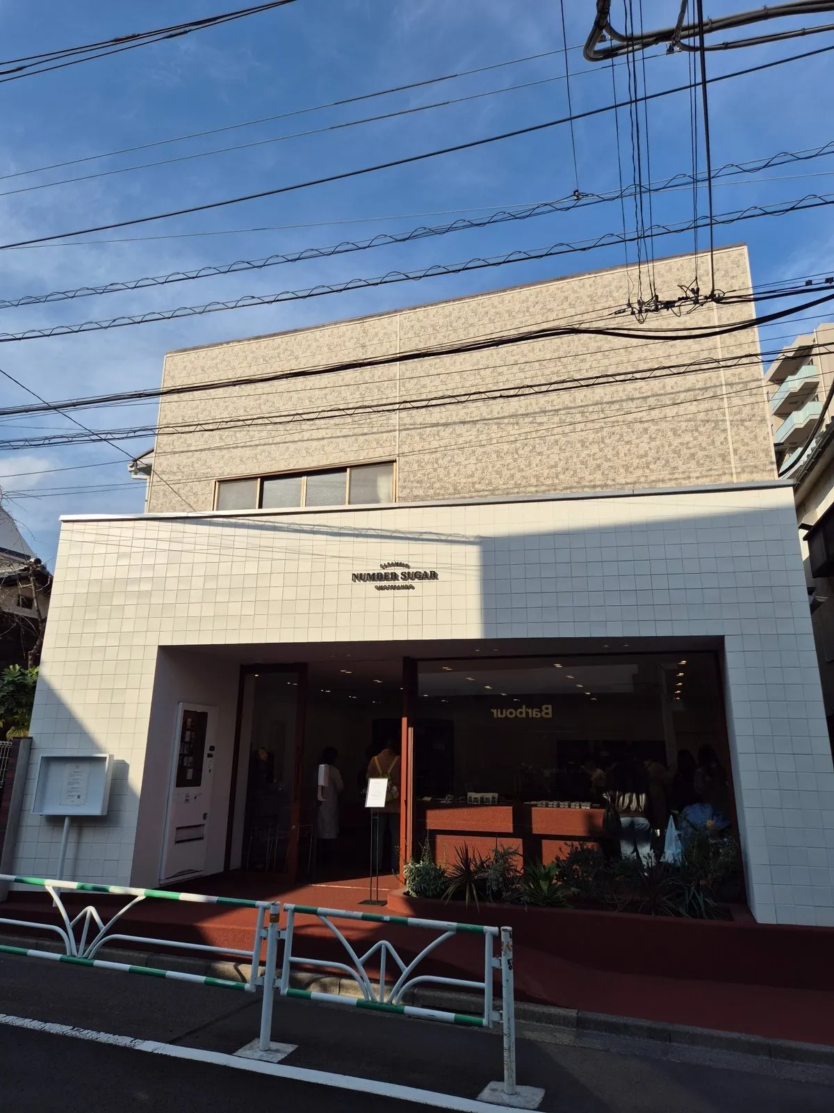 Harajuku, Tokyo'daki hareketli bir sokakta, güneşli bir günde çekilen bu geniş açılı görüntüde, beyaz fayanslarla kaplı modern NUMBER SUGAR dükkanı, cephesindeki HANDMADE CARAMEL yazısıyla ve cam vitrininden belli olan kalabalık iç mekanıyla öne çıkıyor, tepede ise tipik elektrik kabloları mavi gökyüzünü kesiyor.