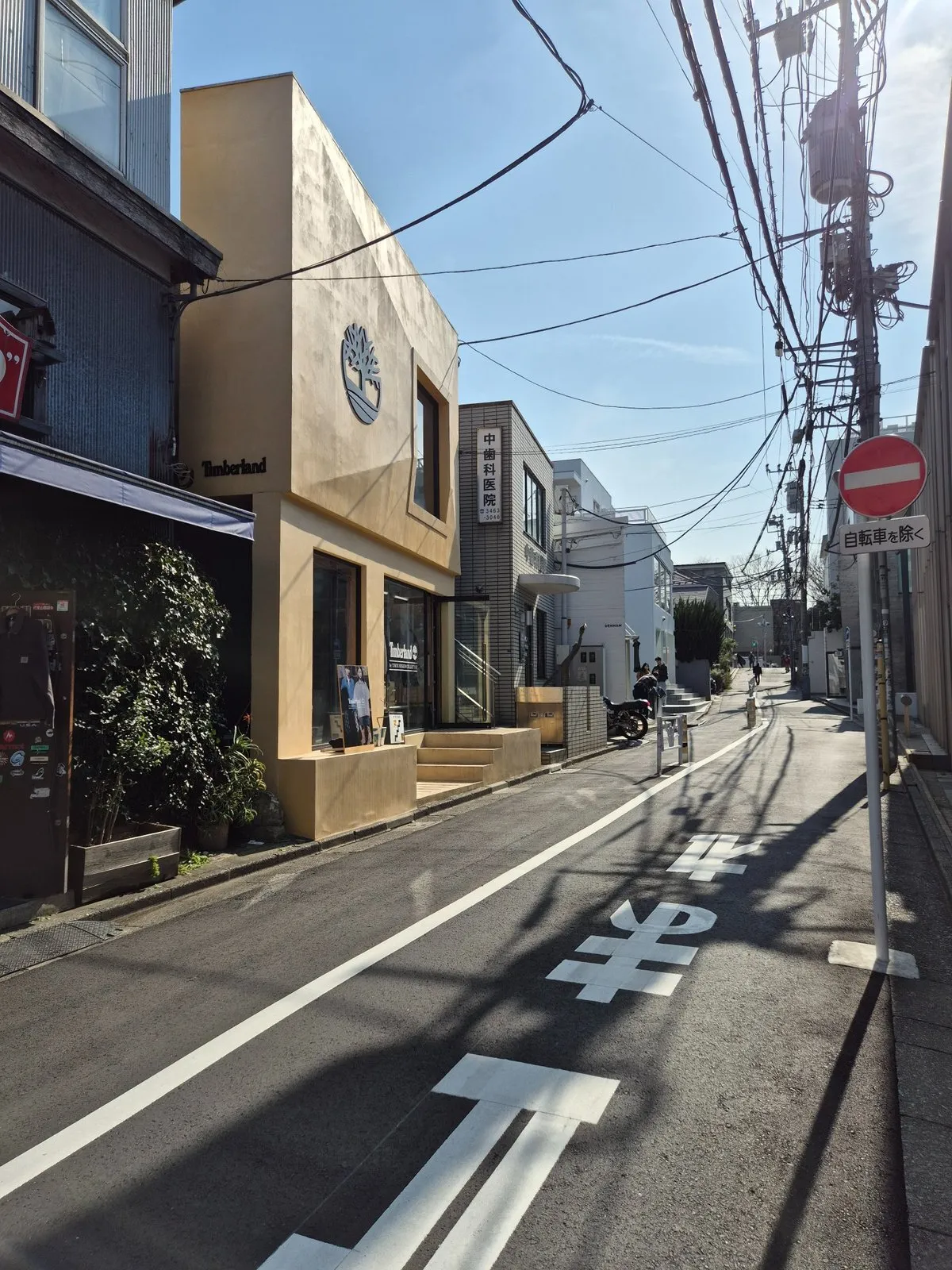 Güneşli bir Harajuku, Tokyo gününde, gökyüzünü saran karmaşık elektrik hatlarının gölgesindeki sakin sokakta, ön planda ağaç logolu bej renkli bir Timberland mağazası ve Japonca diş kliniği tabelalı bitişik bir bina dikkat çekiyor, asfaltta Japonca dur yazısı ve kırmızı yasak işareti ile birlikte kaldırımda park etmiş bir motosiklet de görülüyor.