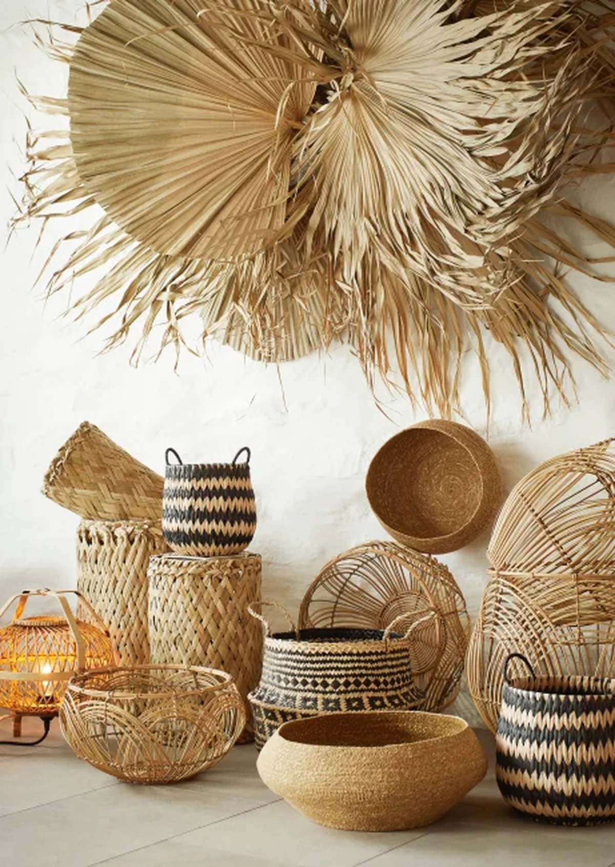 Beyaz bir duvarda, büyük kurutulmuş palmiye yapraklarından oluşan bohem duvar dekorasyonu ile yerde hasır, rattan ve bambu gibi doğal malzemelerden yapılmış, bazıları geometrik desenli çeşitli sepetler ve sıcak ışık veren bir fenerin oluşturduğu doğal ve estetik bir köşe, dış mekan trendine uygun zanaatkar ürünleri sergiliyor.