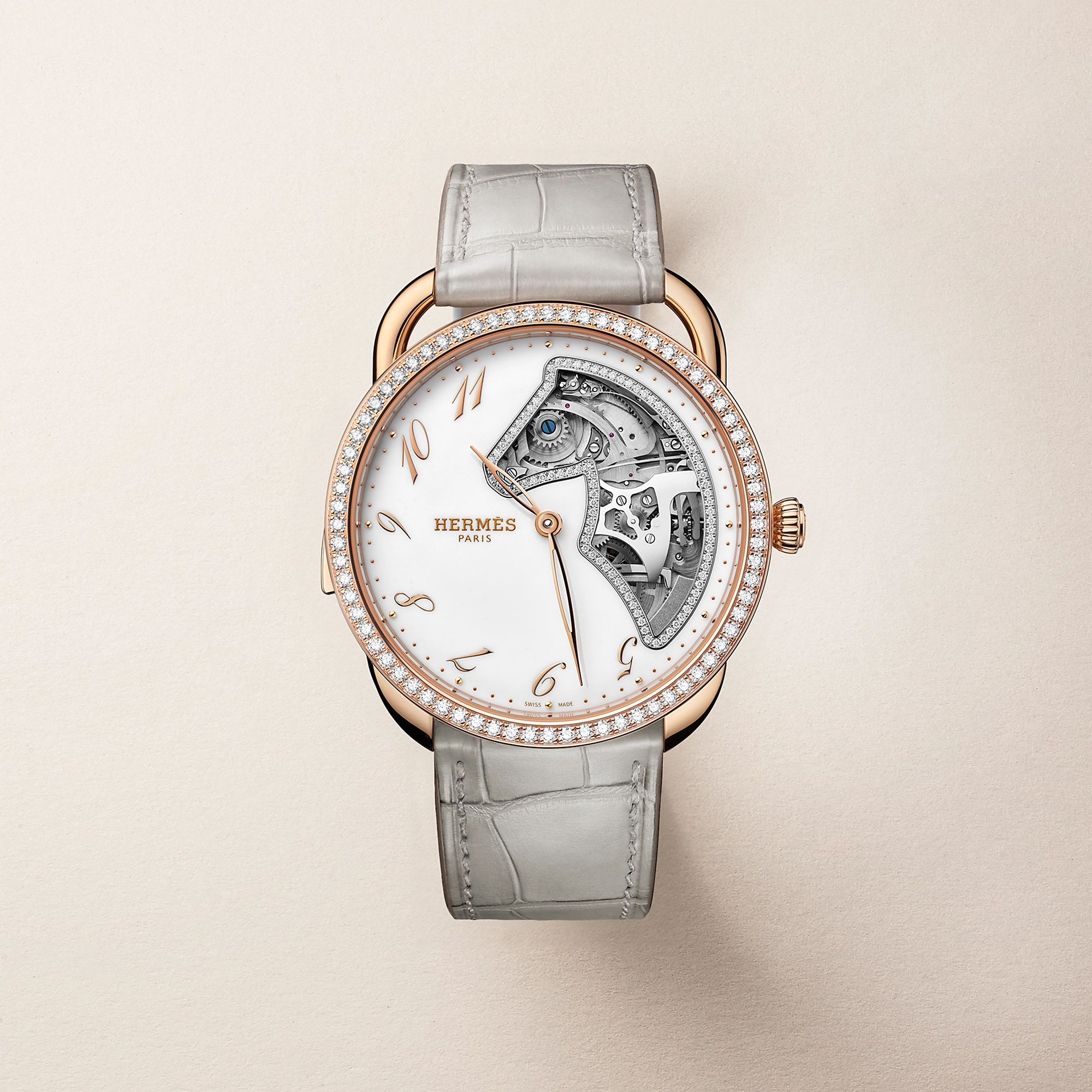 Pırlantalarla çevrili pembe altın kasalı, beyaz kadranında mekanizmanın büyüleyici detaylarını at başı formunda bir kesitle gözler önüne seren ve Watches & Wonders 2026 fuarının öne çıkan kadın saatlerinden biri olan, açık gri timsah desenli kayışa sahip zarif bir Hermès Arceau Samarcande modeli.