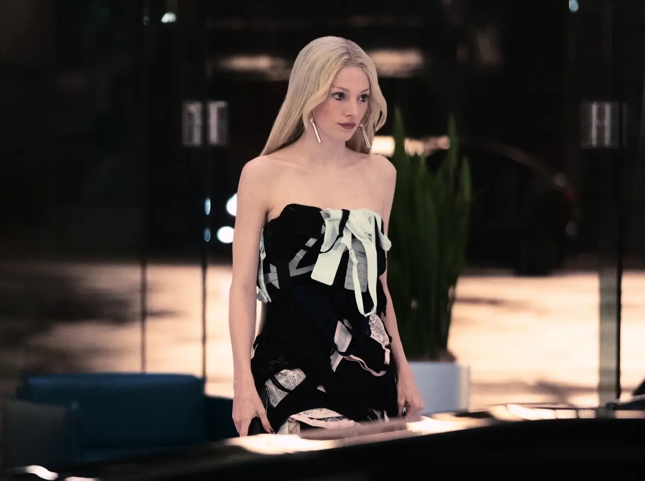 Hunter Schafer, Euphoria dizisinin üçüncü sezonundan olduğu anlaşılan bir fotoğrafta, uzun sarı saçları, dikkat çekici gümüş küpeleri ve siyah ile beyaz iç çamaşırı parçalarından, kurdeleler ve dantellerden özenle tasarlanmış straplez bir elbiseyle, lüks bir mekanda hafifçe yana dönük, düşünceli bir ifadeyle bakıyor.