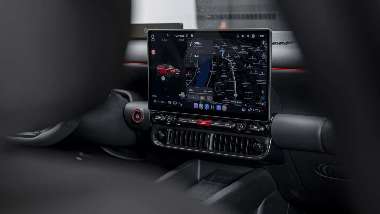 Hyundai Ioniq 3'ün iç mekanından bir görünüm sunan bu fotoğrafta, orta konsolda yer alan geniş dijital multimedya ekranında, kırmızı bir araç simgesiyle birlikte aktif navigasyon haritası ve detaylı yol bilgileri net bir şekilde sergilenmekte, ekranın altında ise fiziksel klima kontrolleri ve havalandırma ızgaraları seçilmektedir.