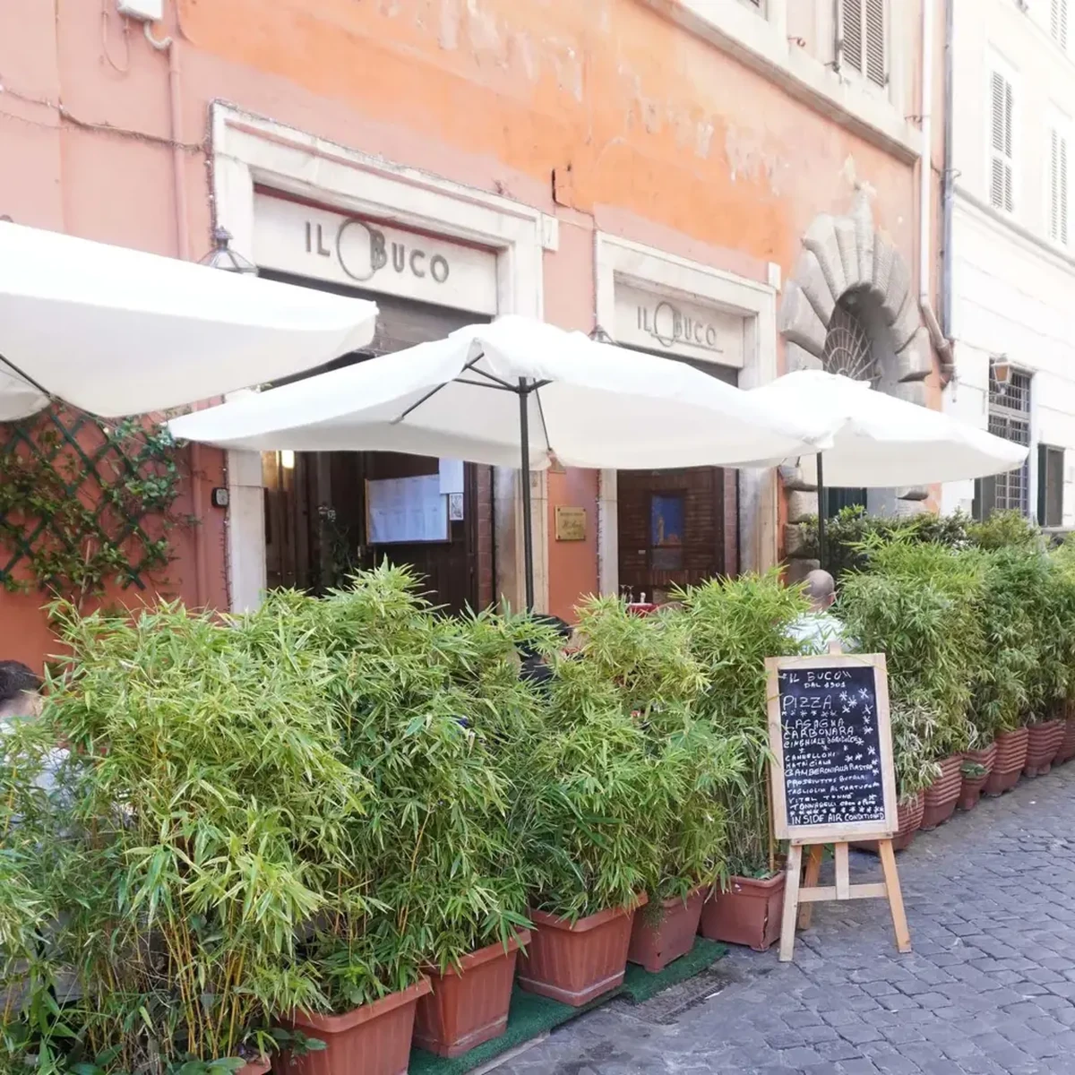 Romantik bir Avrupa sokağında yer alan, ön cephesi turuncu renkli IL BUCO adlı ünlü restoranın, beyaz güneş şemsiyeleri, saksılarda sıralanmış gür yapraklı bitkiler ve kaldırımda duran bir menü tahtasıyla temasına uygun dış cephesi.