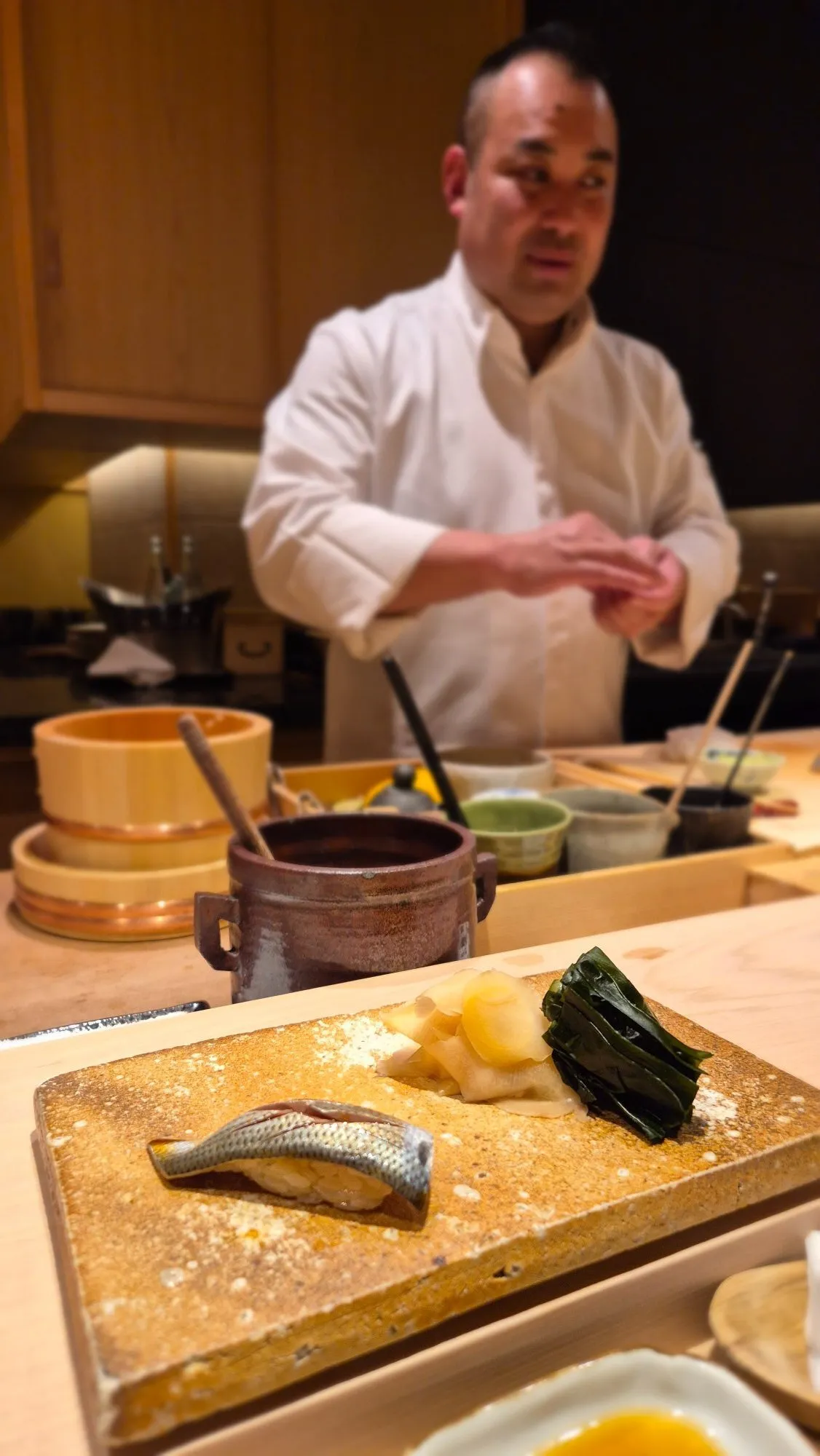Beyaz şef önlüğü giymiş bir aşçı bulanıkça arka planda sushi hazırlarken, ön planda kumlu dokulu bir servis tabağında özenle yerleştirilmiş bir parça nigiri sushi, yanında parlak sarı zencefil turşusu ve koyu yeşil deniz yosunuyla göz alıcı bir sunum sergileniyor.