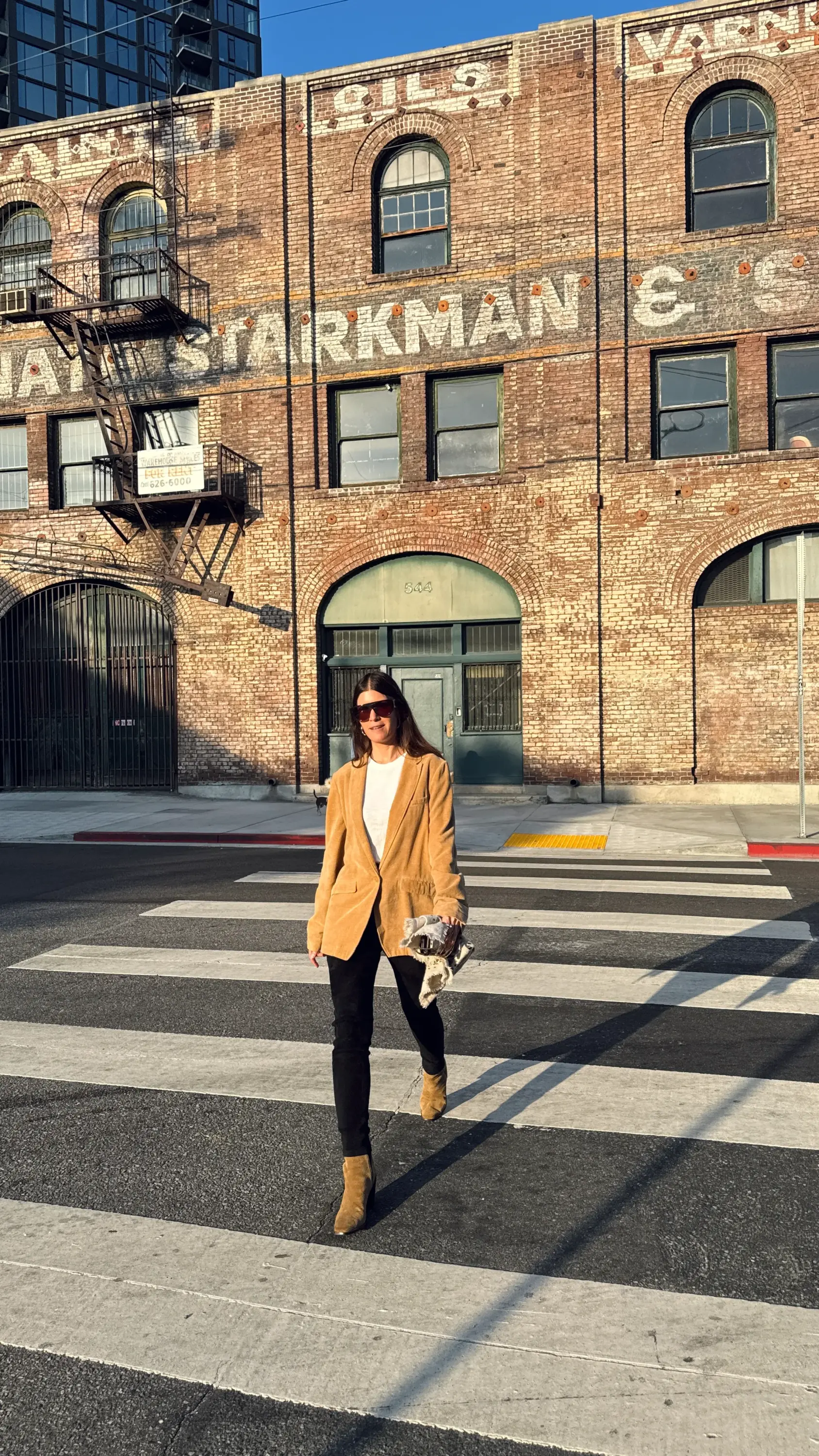 Stylist İpek Ersoy Gelgin, Los Angeles'ın güneşli bir sokağında zebra geçidinden geçerken rahat ama şık bej bir blazer ceket, beyaz tişört, siyah pantolon ve botlarıyla modern stilini yansıtıyor, arkasındaki eski tuğla binalar şehrin estetiğini vurguluyor.