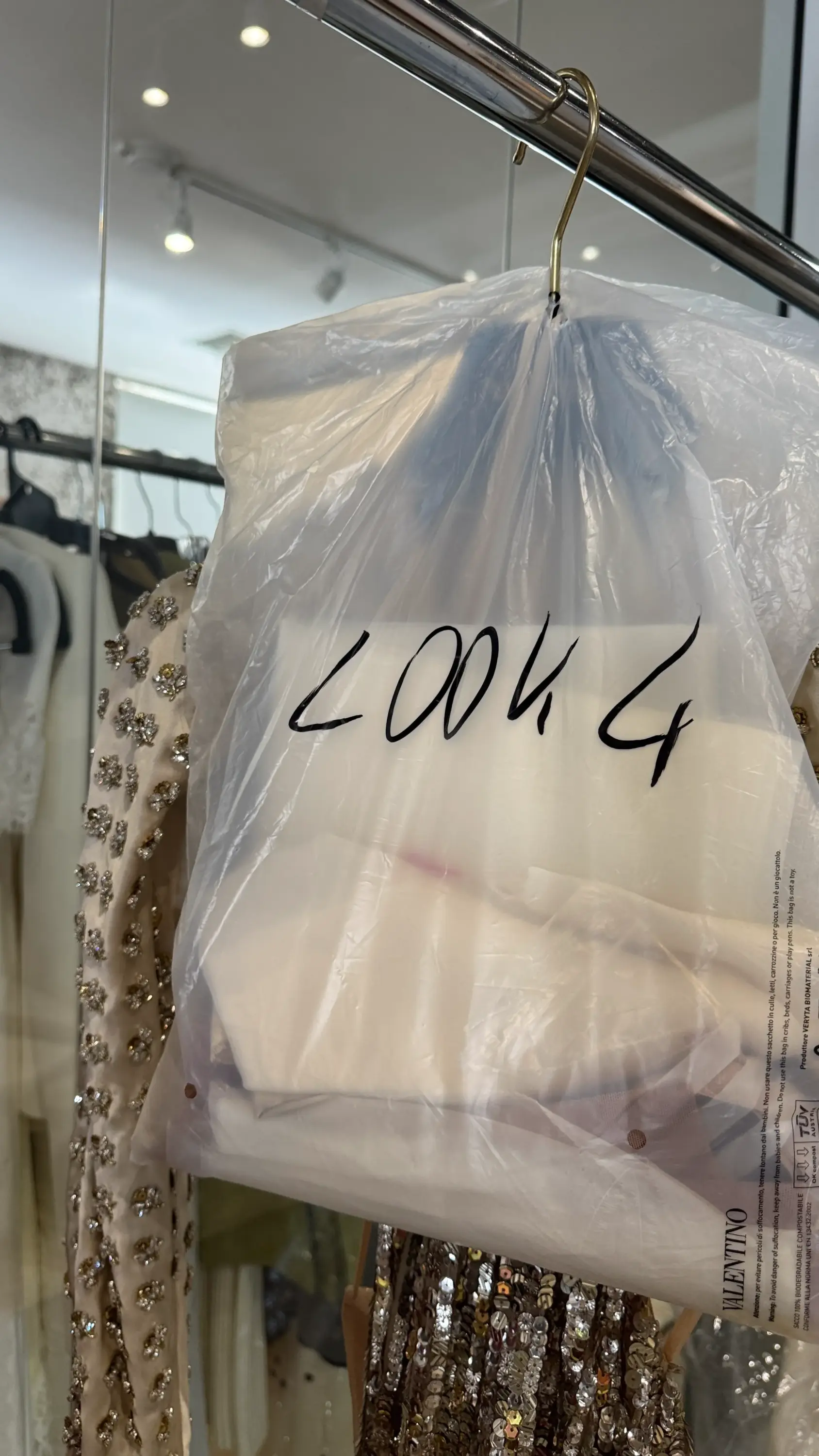 Oscar gecesinin kırmızı halı görünümlerinden birini temsil eden, şeffaf plastik kılıfında siyah kalemle yazılmış LOOK 4 etiketiyle asılı duran ve Valentino markasını taşıyan, parlak işlemeli bir elbise, ünlülerin stil oluşturma sürecinin perde arkasını ve titiz hazırlıklarını vurguluyor.