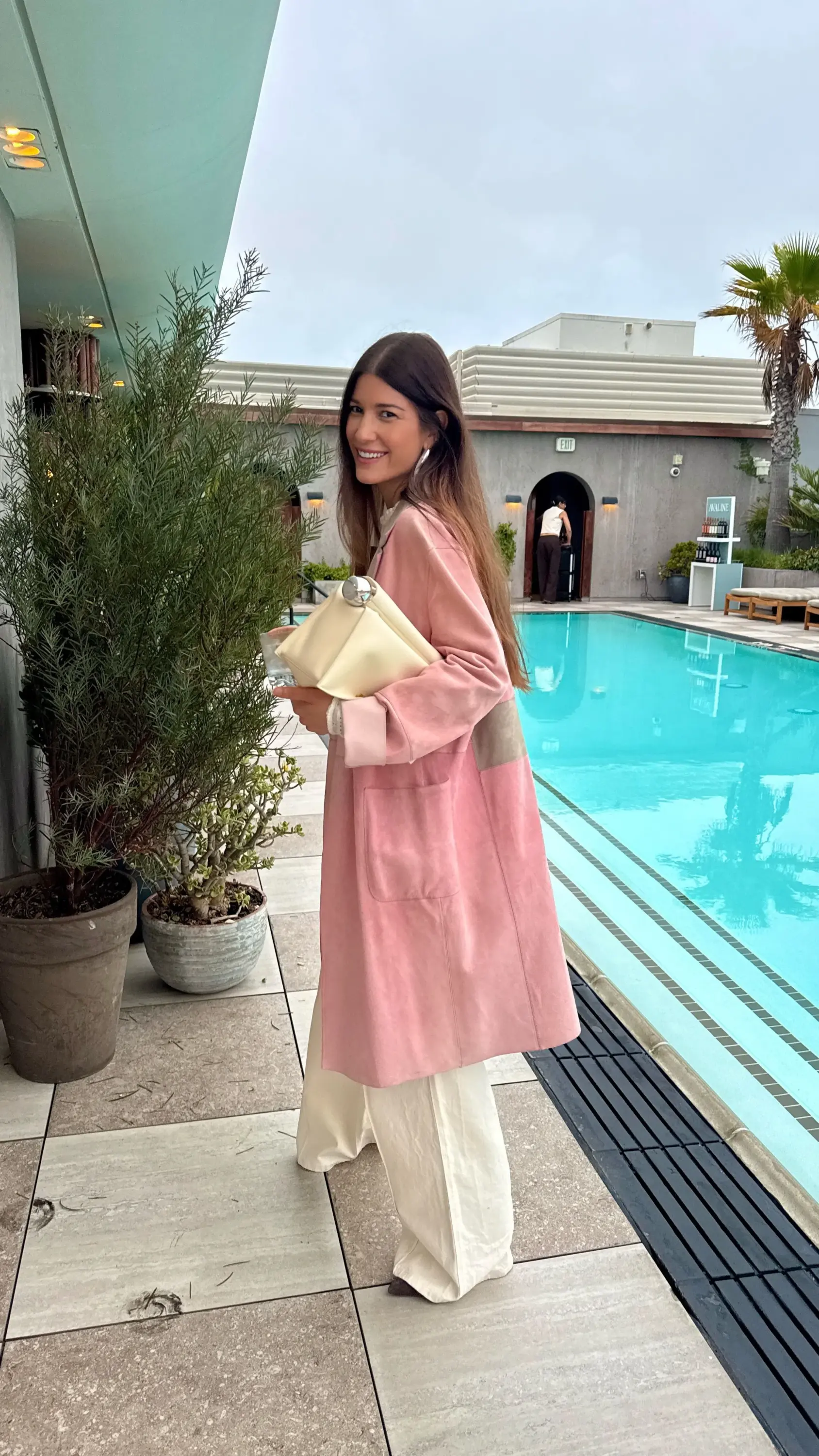 Los Angeles'taki kariyerini sürdüren stylist İpek Ersoy Gelgin, pembe süet ceketi, bol paça krem rengi pantolonu ve zarif krem el çantasıyla havuz kenarında kameraya gülümserken, stil anlayışını yansıtan doğal ve şık bir duruş sergiliyor.