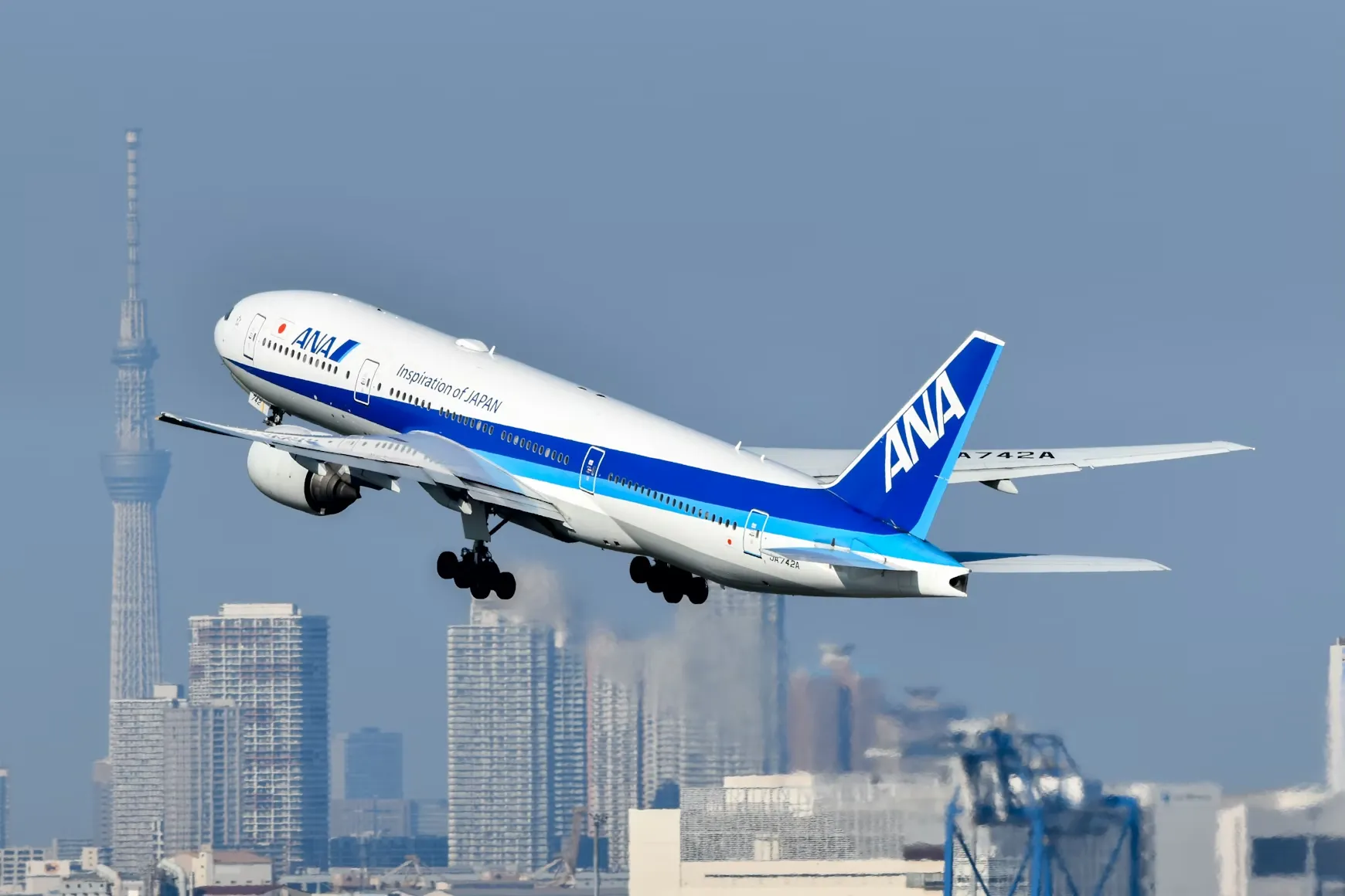 Tokyo Skytree ve şehir silüeti önünde, iniş takımları açık, beyaz ve mavi renkli ANA uçağı, Japonya'ya nasıl uçulur sorusunu yanıtlayan bir hava yolculuğunu temsil ederken gökyüzüne doğru yükseliyor.