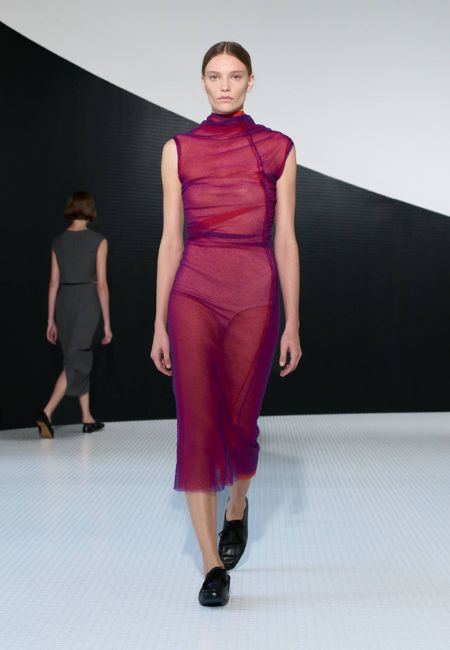 Jil Sander 2026 ilkbahar-yaz moda trendleri sunumundan podyumda yürüyen bir model, transparan mor tonlarında, vücuda oturan, kolsuz midi elbisesi ve siyah bağcıklı ayakkabılarıyla canlı renklerin ve dikkat çekici dokuların önemini vurguluyor.