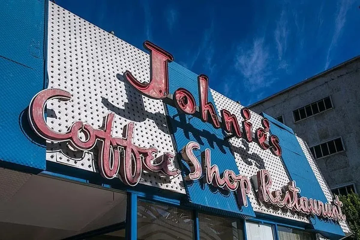 The Big Lebowski ve Reservoir Dogs gibi kült filmlere ev sahipliği yapmış, Los Angeles'ın ikonik Googie mimarisiyle dikkat çeken Johnie's Coffee Shop'un mavi-beyaz çizgili tenteli ve kırmızı neon tabelalı dış cephesi, masmavi gökyüzü altında parlıyor ve sinema tarihine damgasını vuran ünlü restoranlardan biri olarak öne çıkıyor.