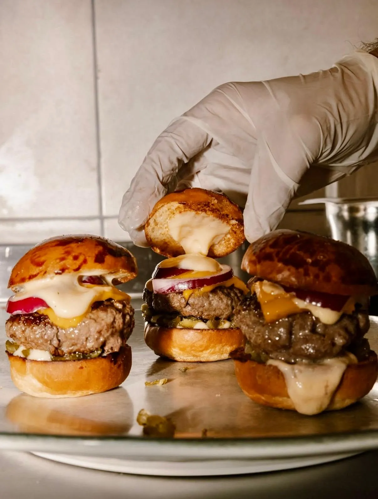 Eldivenli bir elin, eritilmiş peynir ve sosla kaplı mini burgerin üst ekmeğini yerleştirdiği anı gösteren bu yakından çekim fotoğrafta, Jordi Yeniköy'ün lezzetli burgerlerinden üç tanesi parlak bir yüzey üzerinde iştah açıcı bir şekilde sergileniyor.