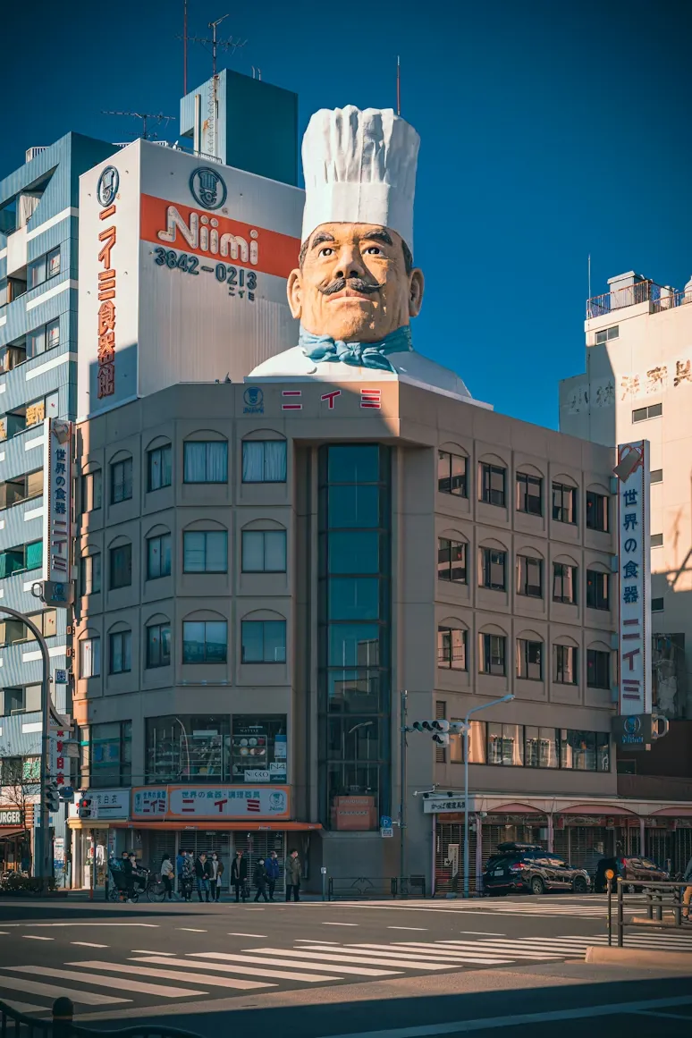Tokyo'daki ünlü Kappabashi mutfak eşyaları bölgesinde yer alan Niimi mağazasının çatısında, devasa beyaz şef şapkası ve mavi fularıyla dikkat çeken, güneşli bir gökyüzü altında duran bir aşçı heykeli ve kalabalık bir cadde görünüyor.