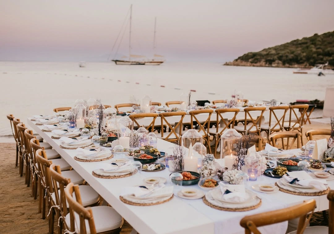 Gün batımının romantik ışıkları altında, Bodrum’un kumlu sahilinde, zarif ahşap sandalyeler ve denize özgü dekorasyonlarla bezenmiş uzun beyaz bir masanın, misafirlere Türk Rivierası’nda unutulmaz bir düğün yemeği deneyimi sunduğu büyüleyici bir akşamüstü manzarası.