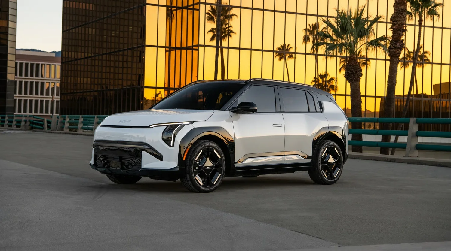 Altın sarısı yansımalara sahip modern cam bir binanın ve palmiye ağaçlarının önünde park edilmiş, beyaz renkte ve siyah detaylara sahip şık bir Kia EV3 elektrikli SUV, minimalist ve fütüristik tasarımıyla dikkat çekiyor.