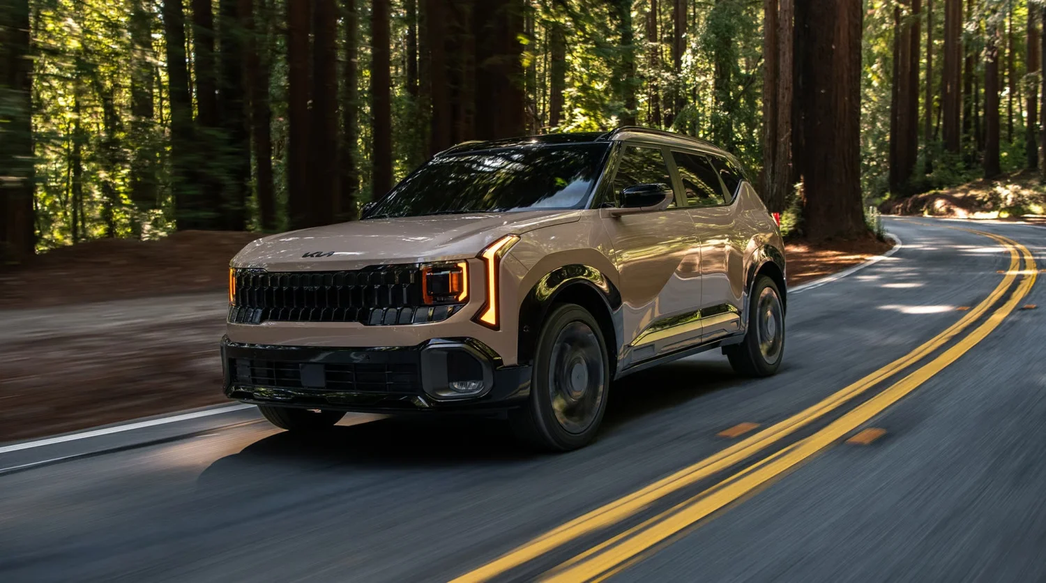 Bej renkli Kia Seltos elektrikli SUV, ormanlık alanda uzanan virajlı asfalt bir yolda, ağaçların gölgesinden süzülen güneş ışıkları altında yol alıyor; belirgin ön ızgarası ve parlak gündüz farları dikkat çekiyor.