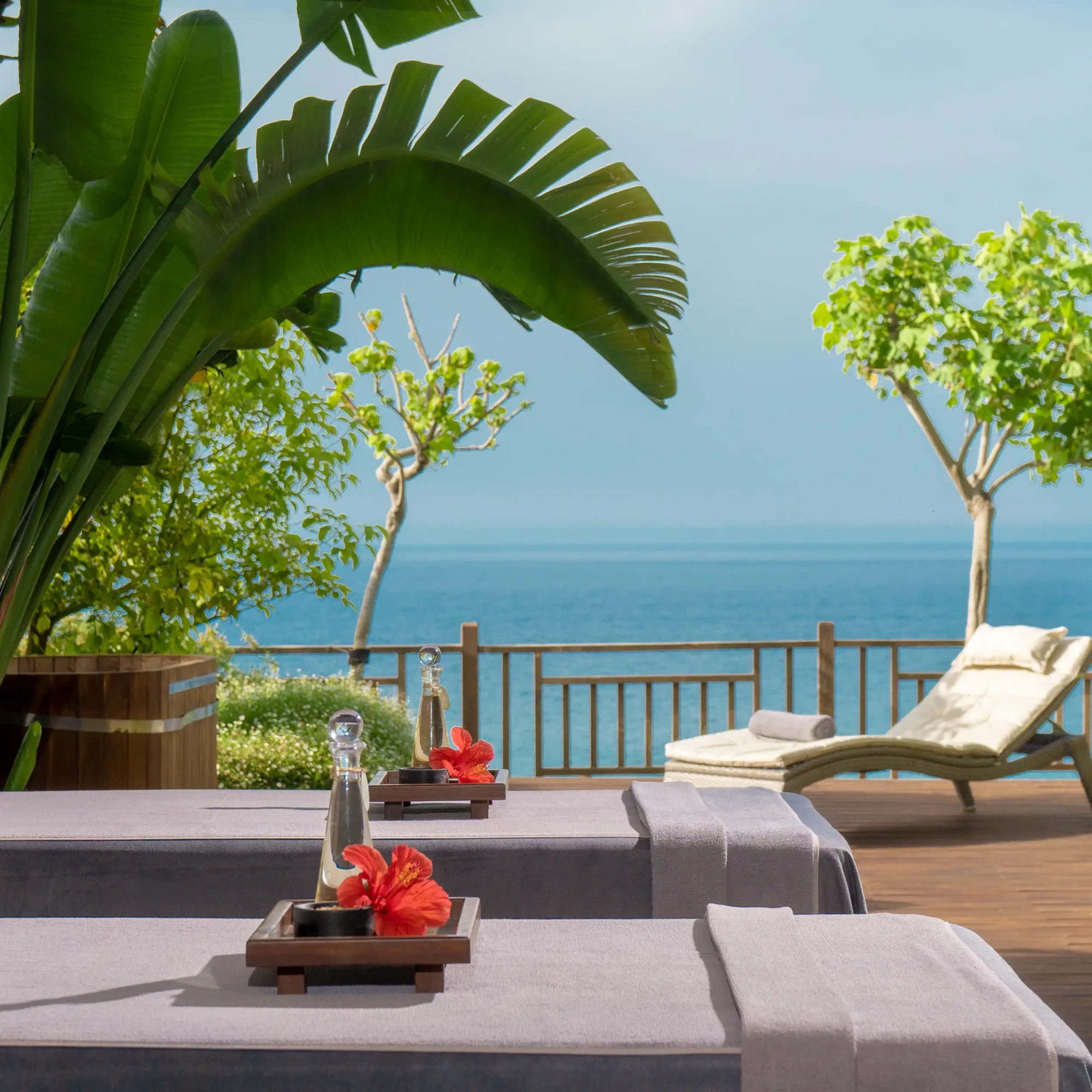 Kıbrıs'taki Elexus Hotel Resort & Spa'nın huzurlu deniz manzaralı spa alanında, ahşap bir teras üzerinde yer alan masaj yatakları, masaj yağı ve canlı kırmızı hibiskus çiçekleriyle süslenmiş olup, tropikal bitkilerin gölgesinde lüks bir dinlenme vadediyor.
