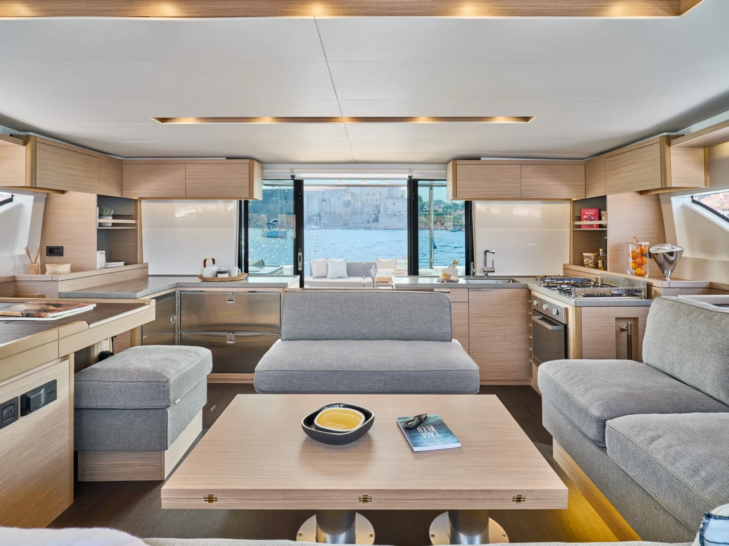 Lagoon 51 model yatın modern ve aydınlık iç mekanında, açık renk ahşap mobilyalarla döşenmiş geniş salon ve tam donanımlı mutfak alanı, büyük cam pencerelerden görünen masmavi deniz manzarasıyla lüks bir yaşam hissi sunuyor.